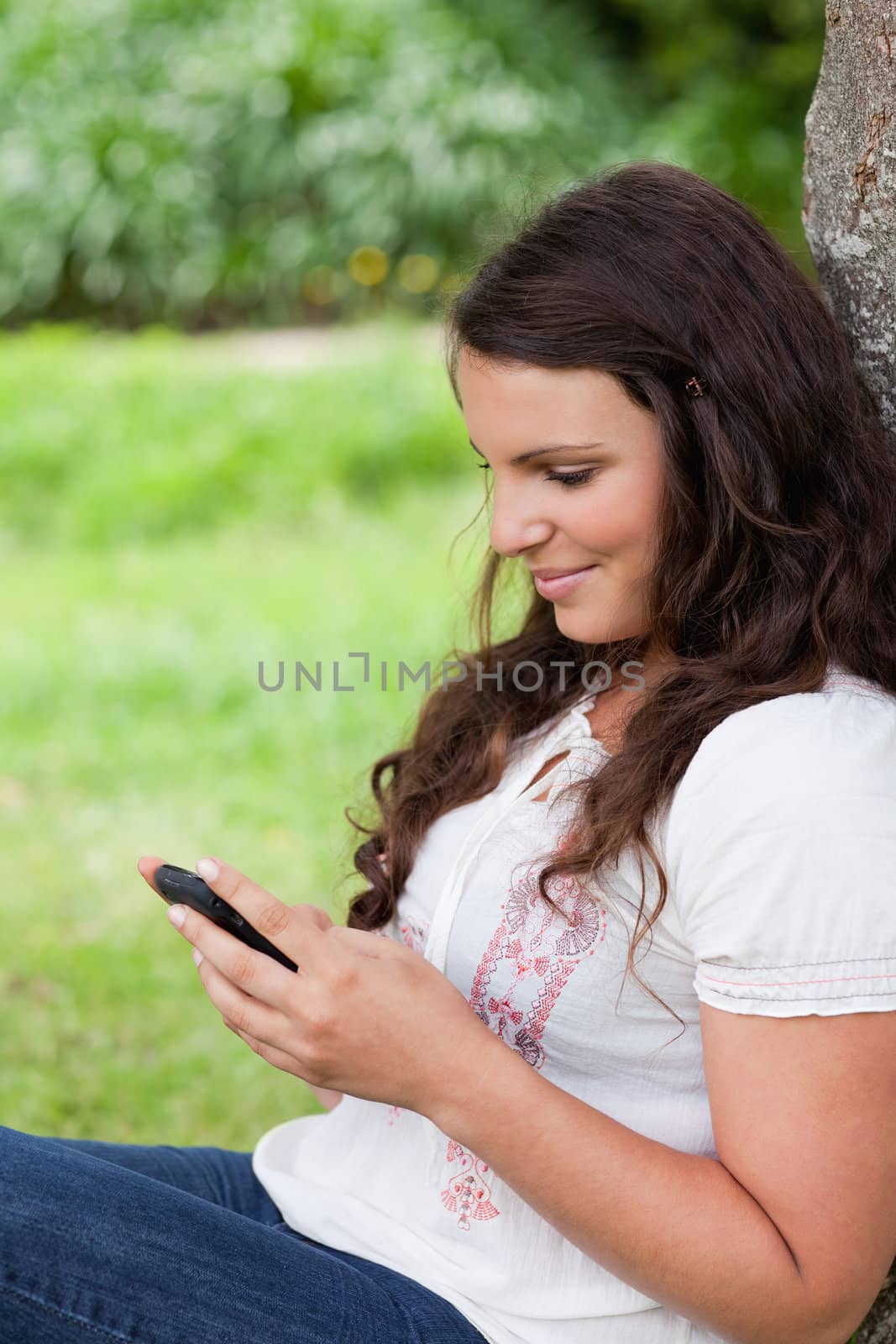 Young relaxed woman sending a text while sitting against a tree by Wavebreakmedia