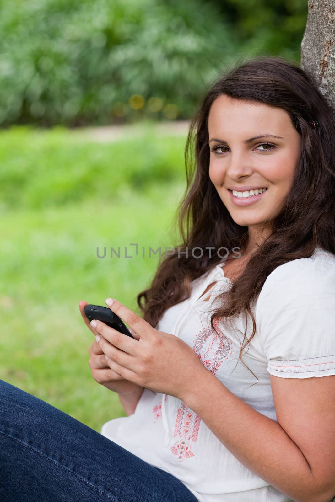 Young smiling woman leaning against a tree while sending a text with her cellphone