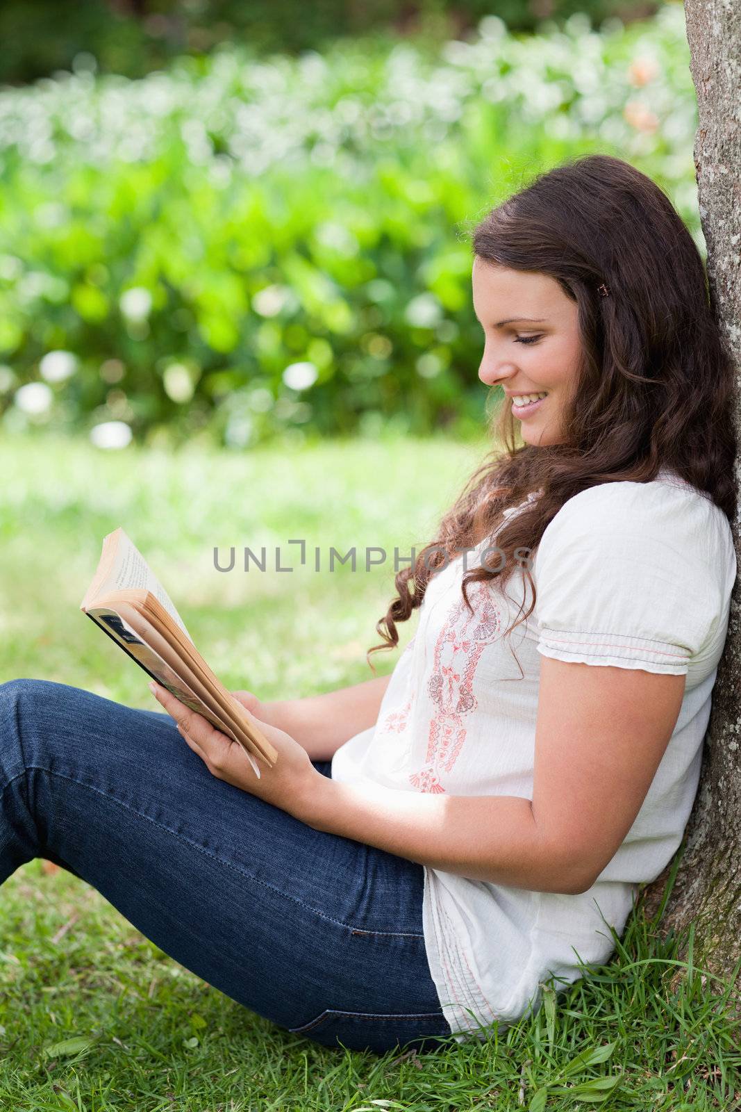 Young smiling girl reading a book while sitting against a tree in the countryside
