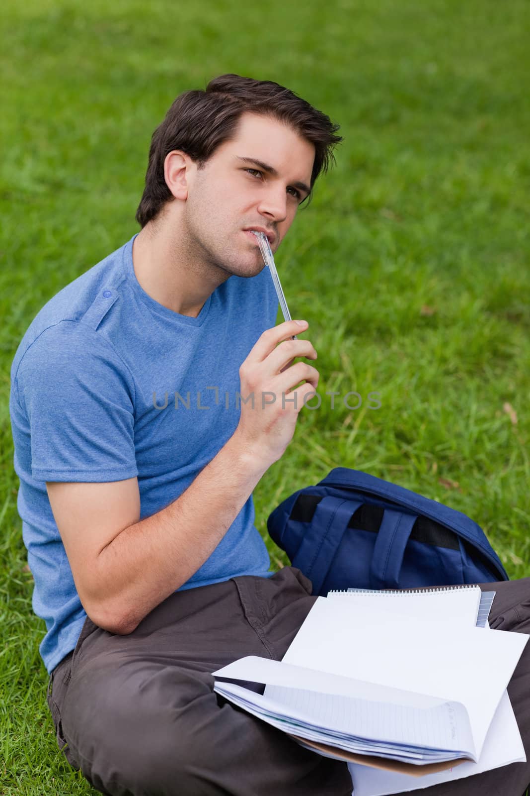 Young thoughtful man holding his ballpoint pen while sitting on the grass in a park