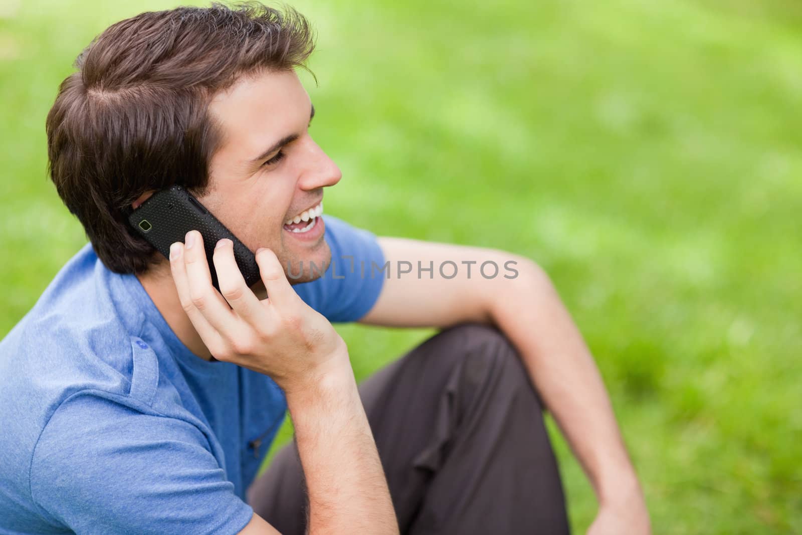 Young man talking on the phone while laughing and sitting on the grass in a park