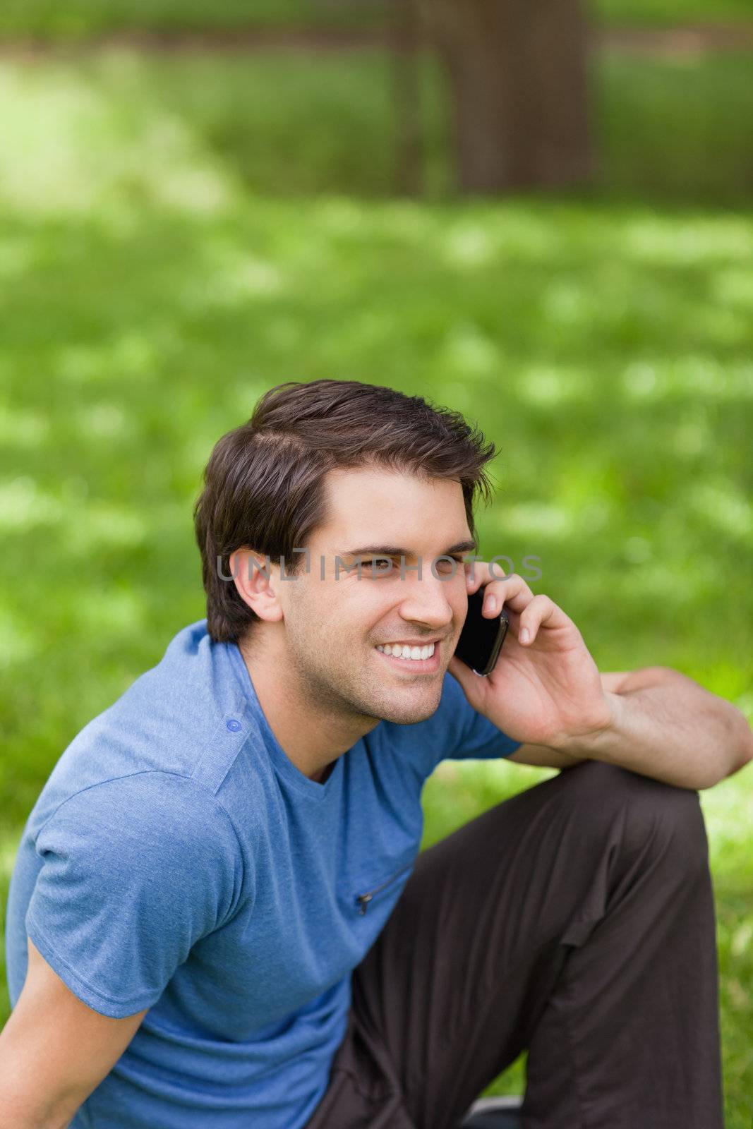 Young smiling man talking on the phone while sitting on the grass in a public garden