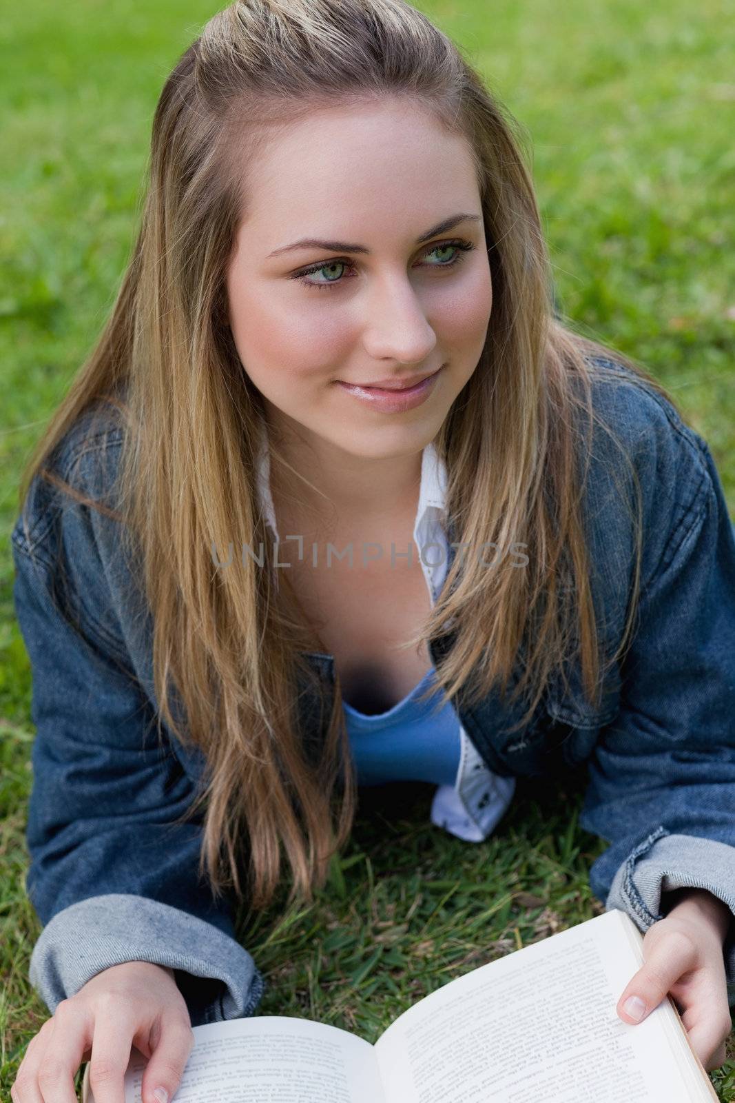 Young smiling girl looking away while lying on the grass in a park and reading