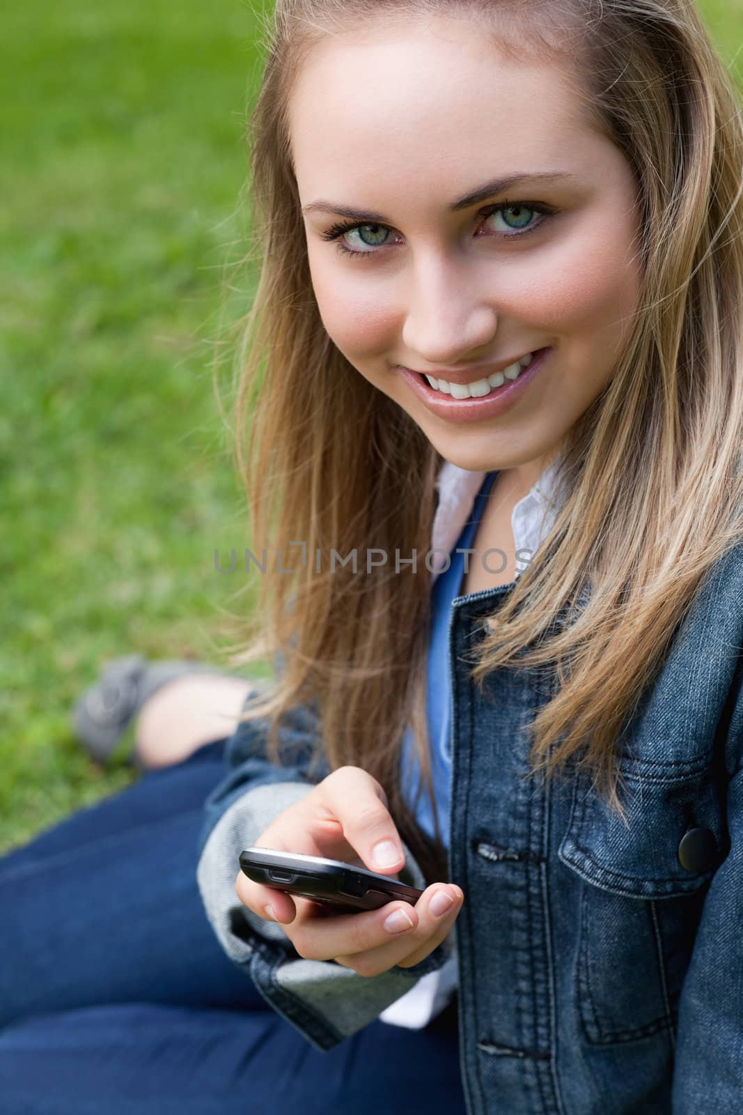 Young blonde girl sitting on the grass in a park while sending a text with her cellphone