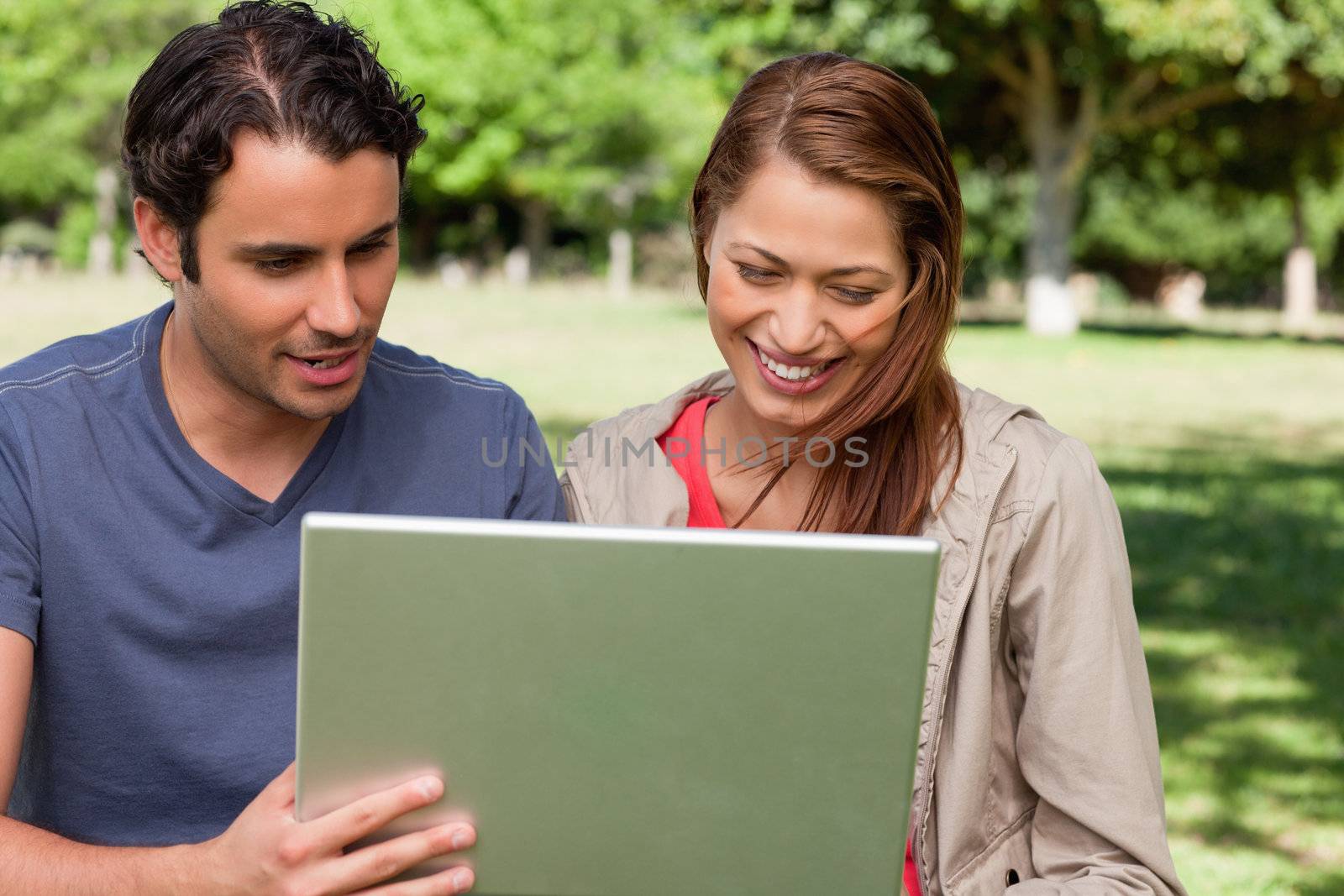 Man holds a tablet as he and his friend watch something on its screen while sitting in a bright park