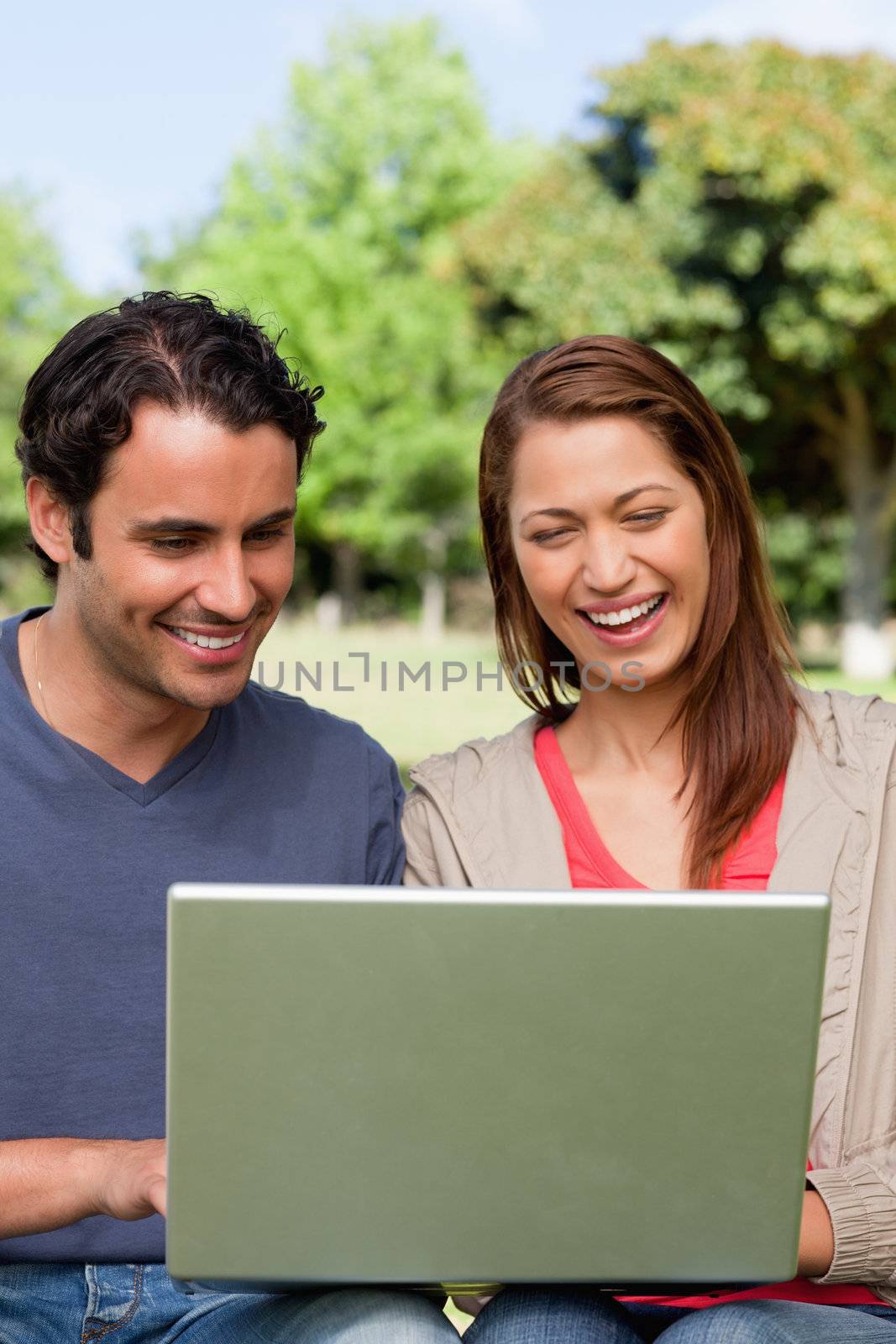 Two friends laughing as they watch something on a tablet while sitting together in a bright area surrounded by trees