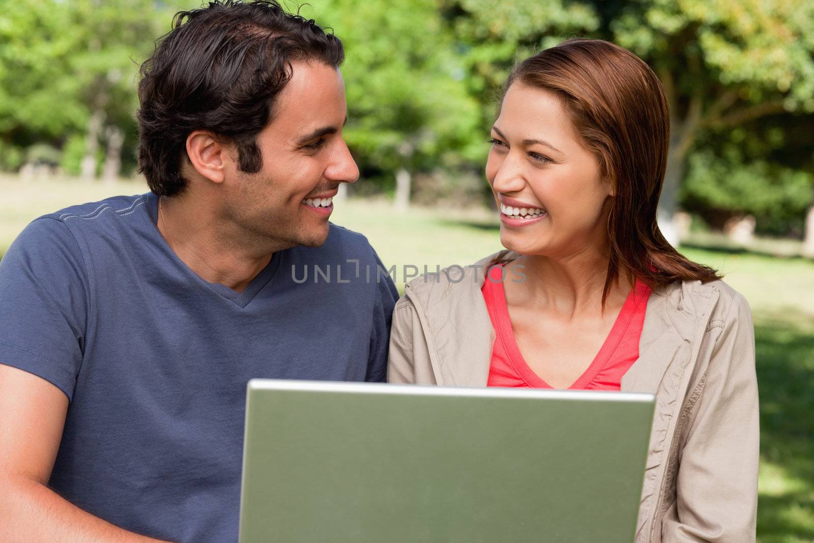 Two friends joyfully smiling as they look at each other while holding a tablet in a park on a sunny day