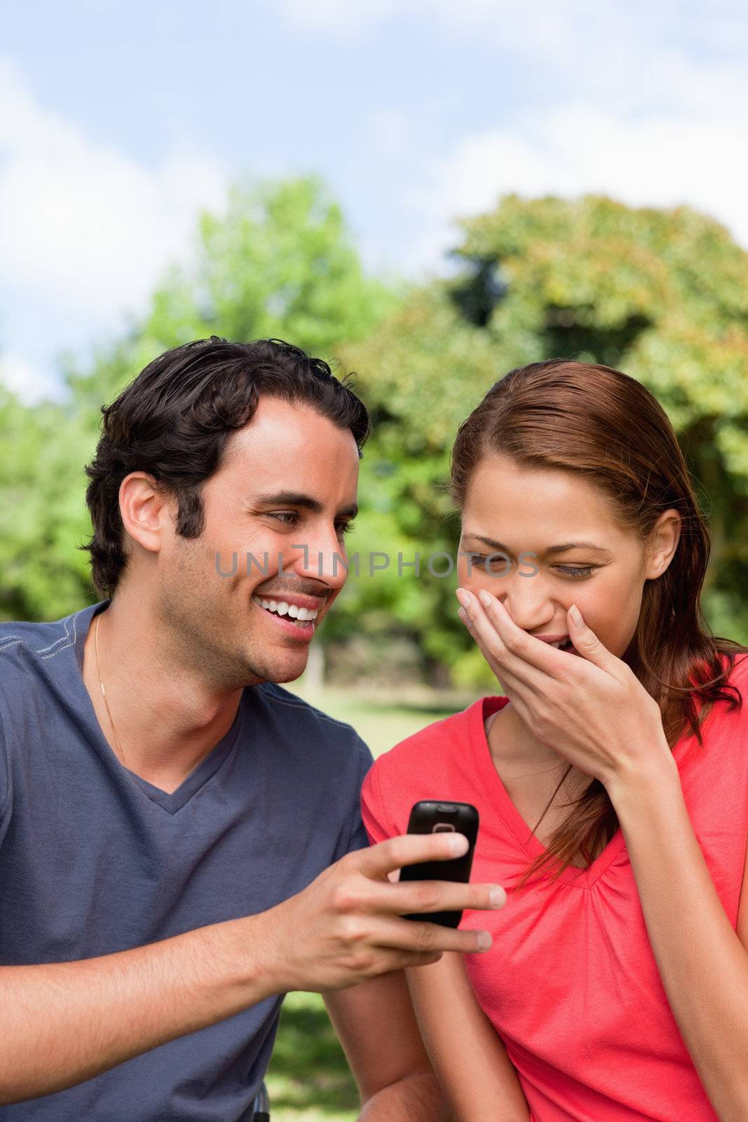 Woman covering her mouth as she is laughing at something being shown to her on her friend's mobile phone in a bright parkland