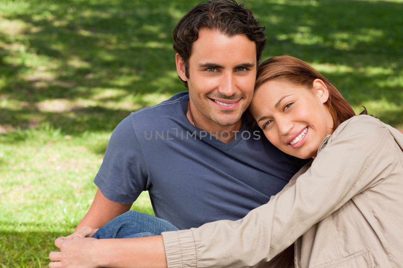 Man looking towards the sky while smiling as his friend rests his rests her head on his shoulder while they lie sit o the grass