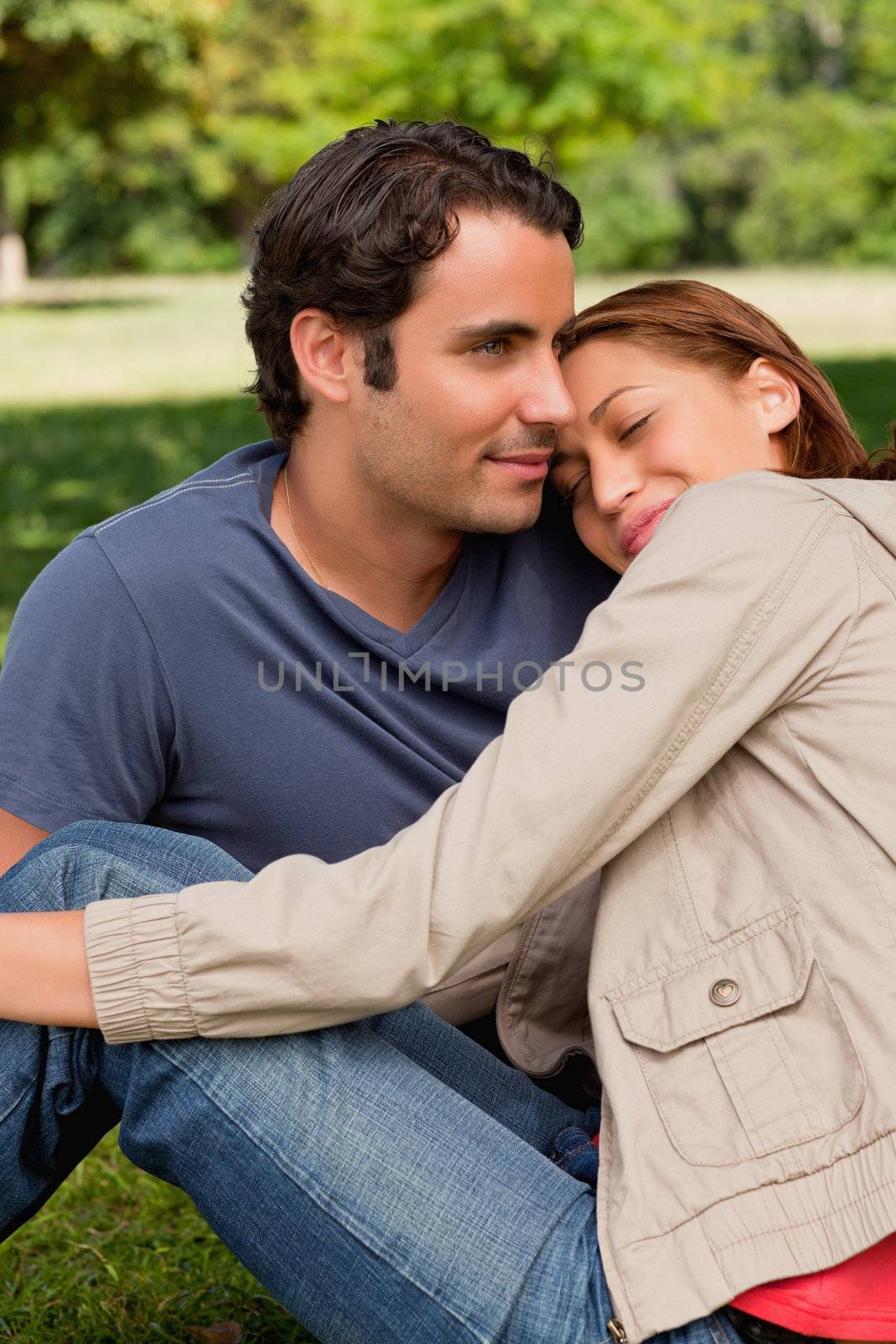 Man looking into the distance as his friend is resting her head on his shoulders with her eyes closed while they both sit on the grass