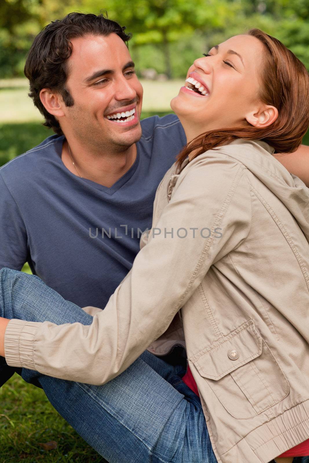 Two friends gleefully laughing as they are sitting next to each other on the ground in a shaded area of a park