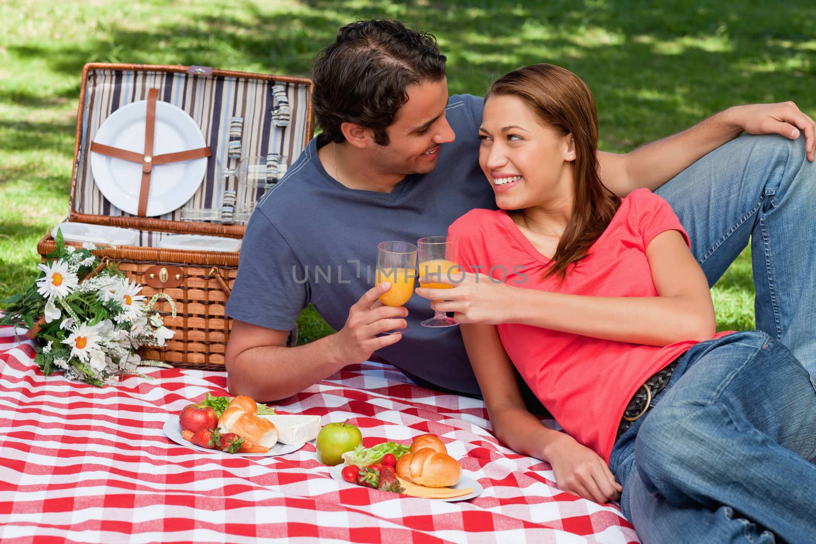 Two friends touching glasses while looking at each other as they lie together on a blanket with picnic food