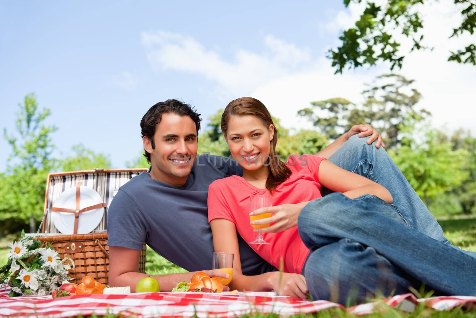 Two smiling friends looking ahead while they hold glasses as they lie on a blanket with a view of the sky in the background