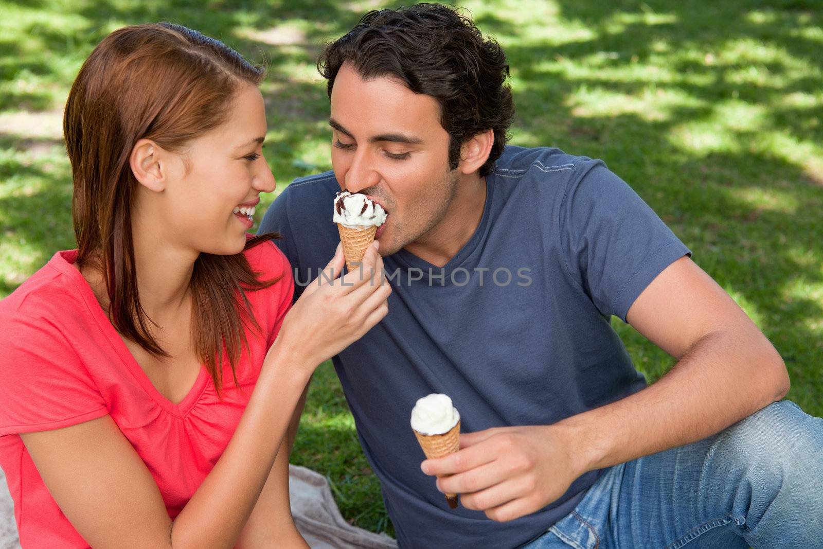 Smiling woman feeding her friend an ice cream cone as they sit next to each other on the grass