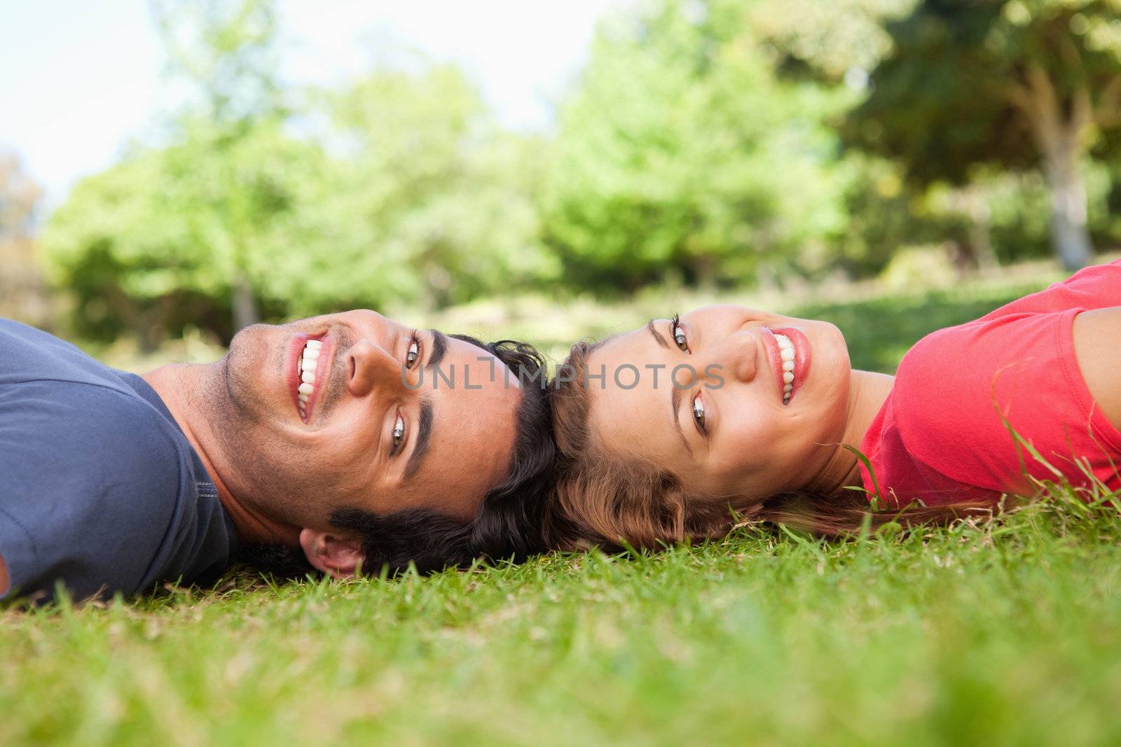 Two friends smiling while looking towards the side as they lie head to head on the grass