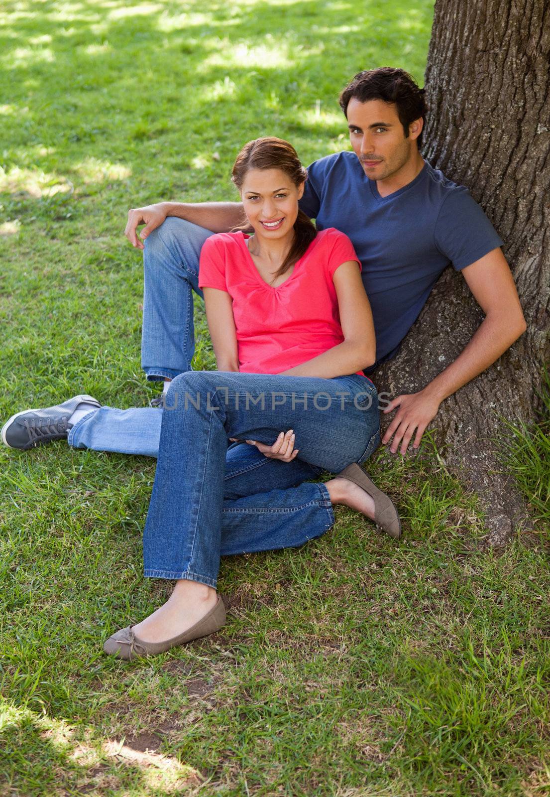Two smiling friends looking towards the side while sitting together against the trunk of a tree