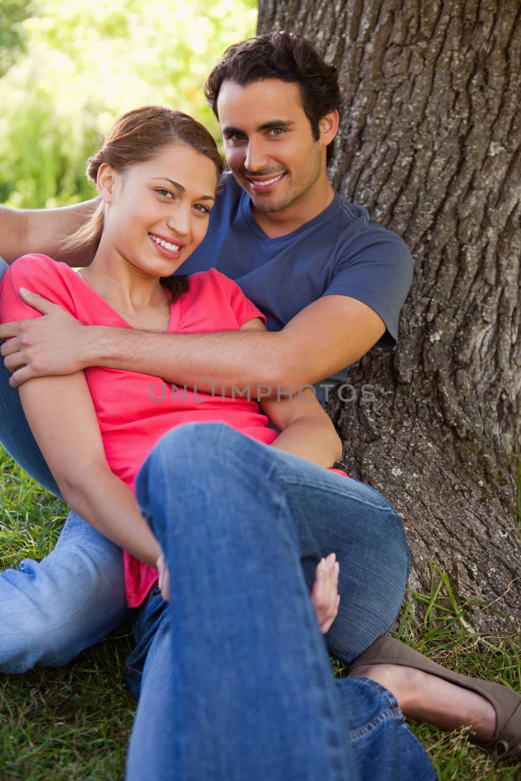 Man looking straight ahead as he holds his smiling friend while they sit together against the trunk of a tree