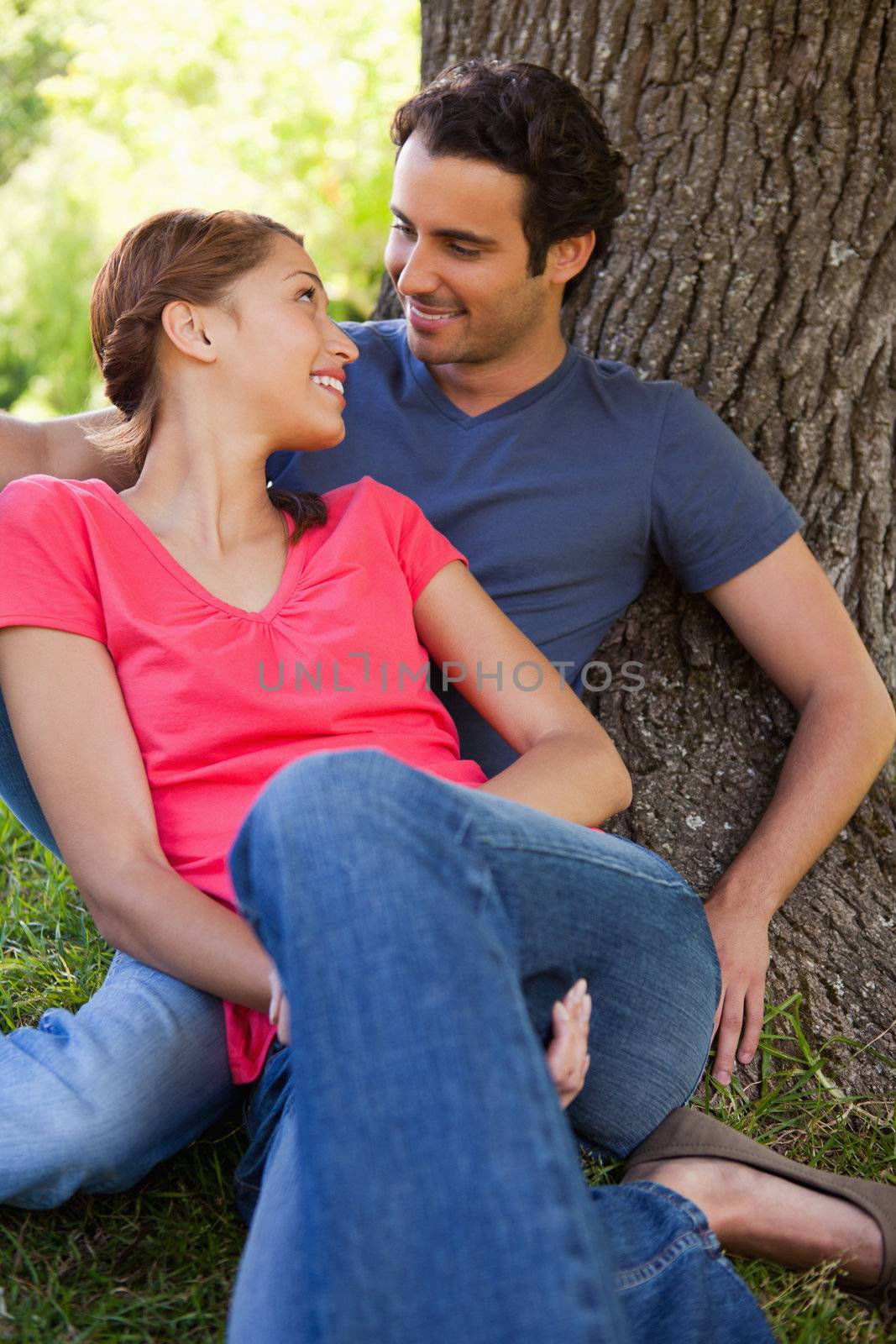 Two friends smiling while looking at each other as they sit together against the trunk of a tree