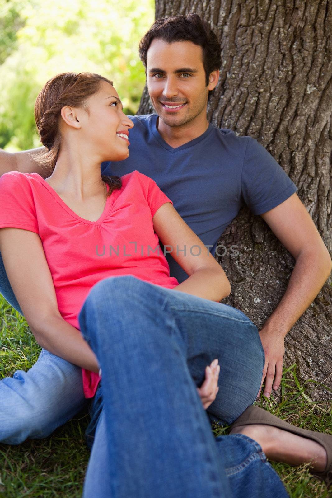 Woman smiling while looking at her friend as they sit together against the trunk of a tree