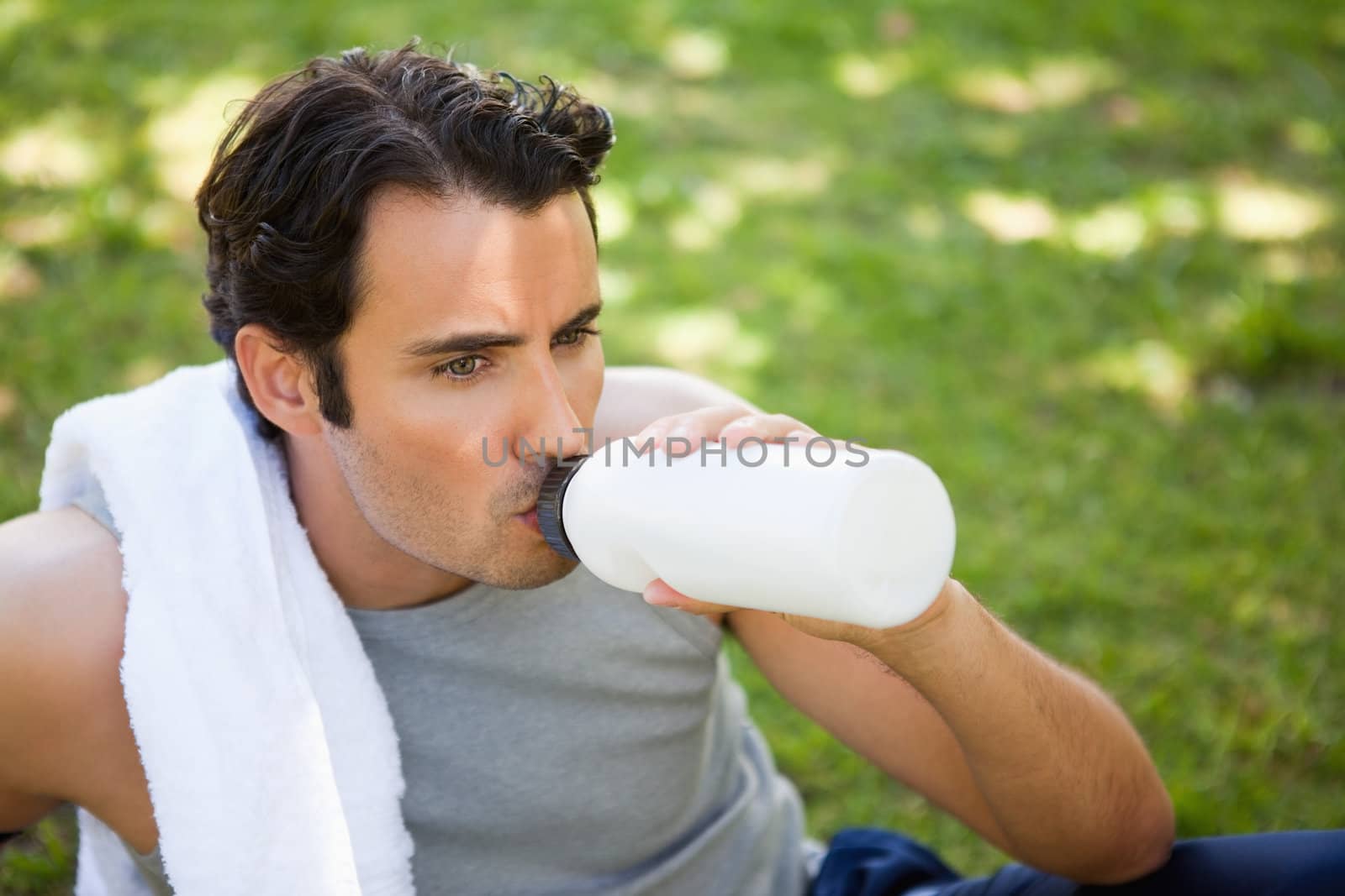 Man with a white towel on his shoulder drinking from a sports bottle while sitting on the grass