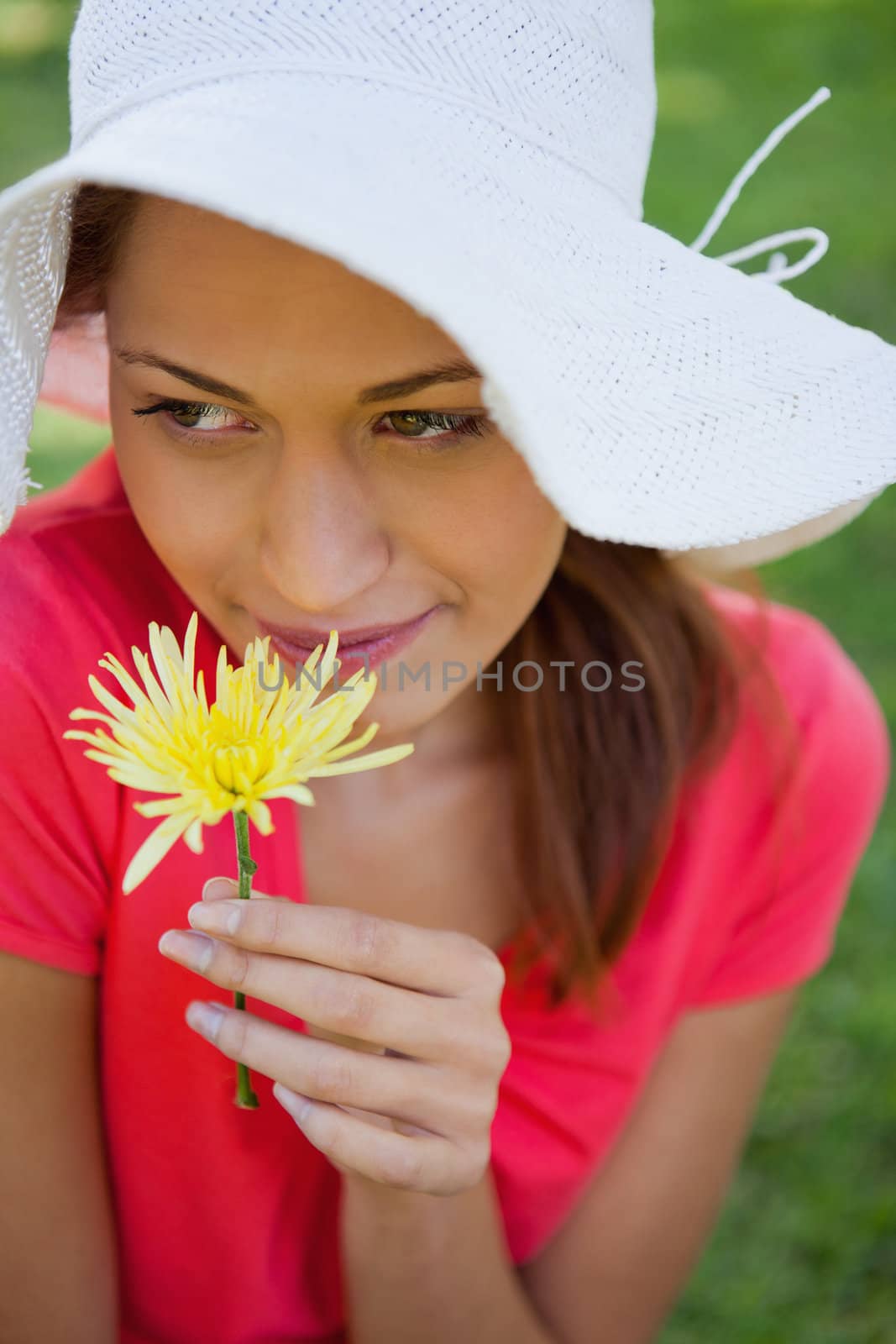 Woman wearing a white hat while smelling a yellow flower while looking towards the side, with grass in the background