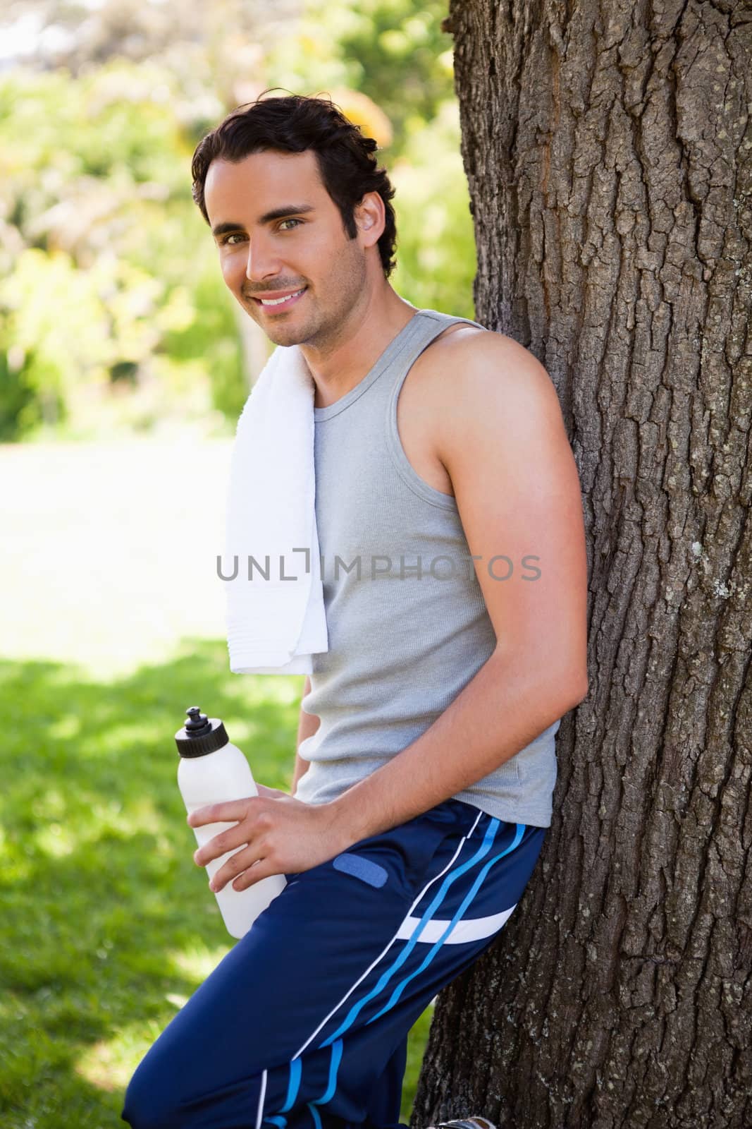 Smiling man with a towel on his shoulder looking towards side while holding a water bottle and resting against the trunk of a tree