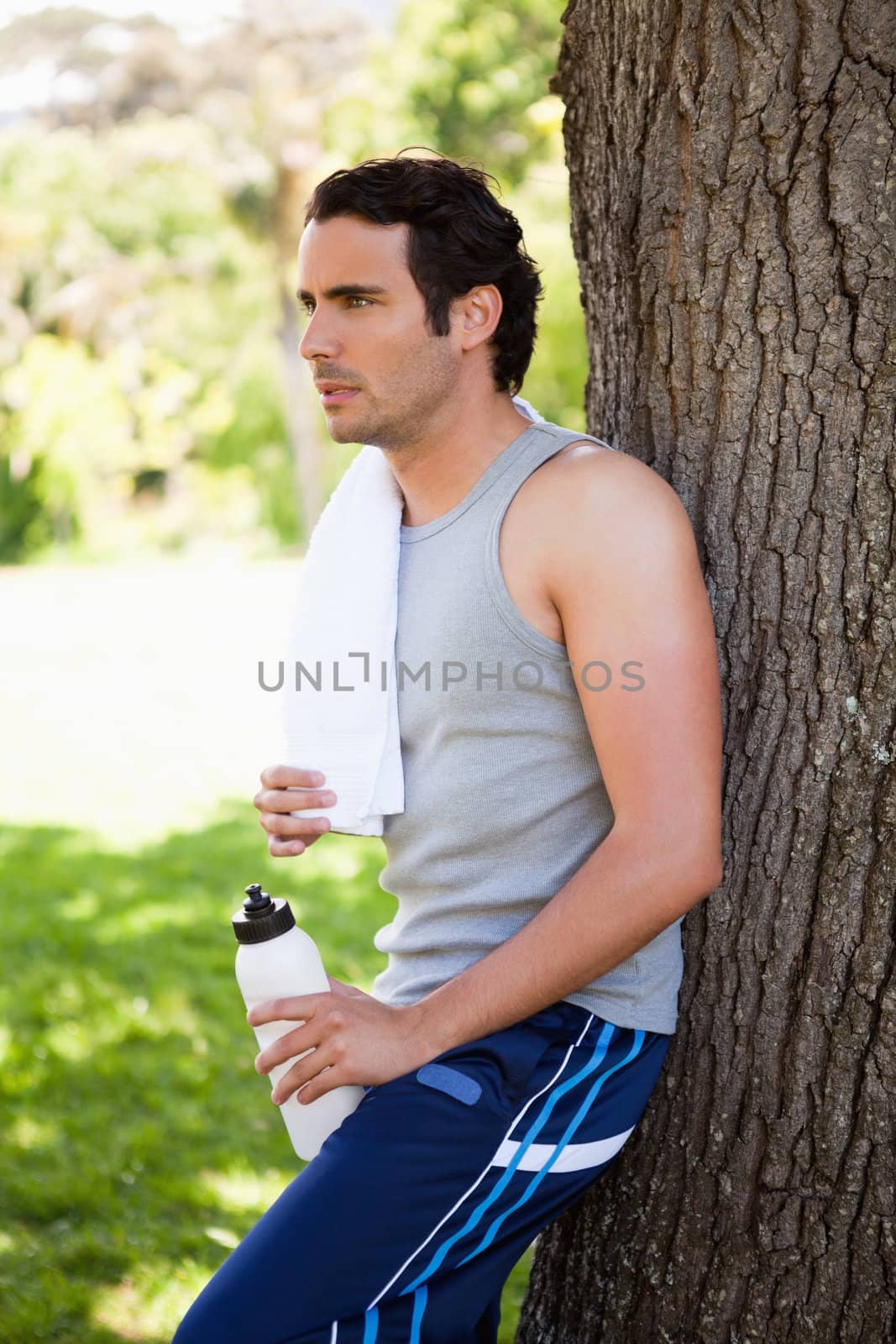 Man with a white towel on his shoulder, holding a sports bottle while leaning against the trunk of a tree