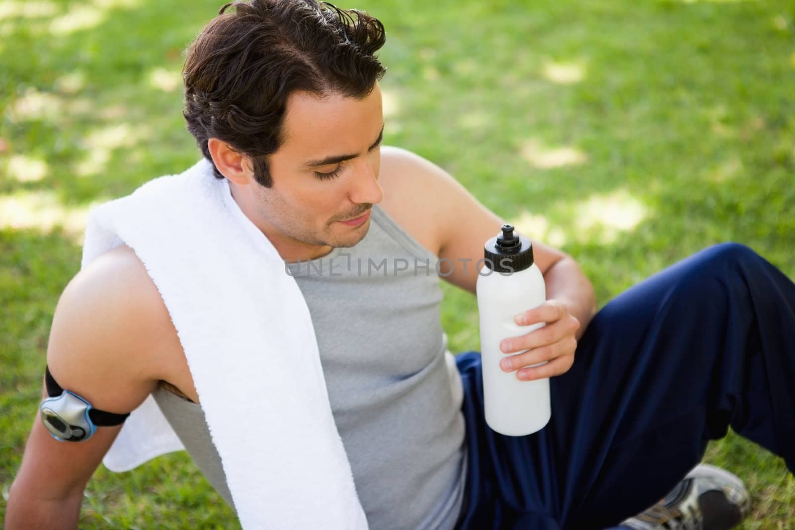 Man with a white towel resting on his shoulder looking at a sports bottle while he is sitting on the grass
