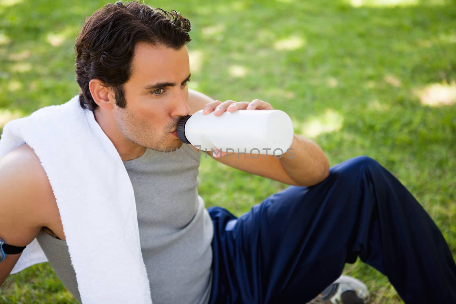 Man with a white towel on his shoulder looking straight ahead while drinking from sports bottle as he sits on the grass
