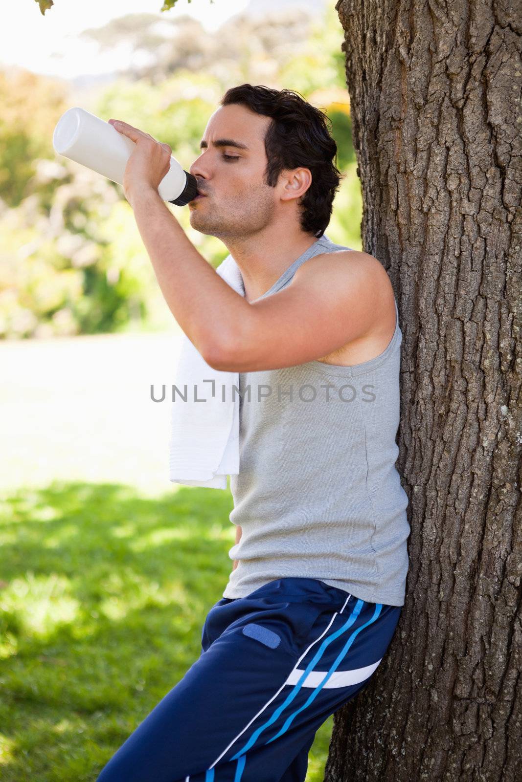Man with a towel on his shoulder drinking from a water bottle while resting against the trunk of a tree