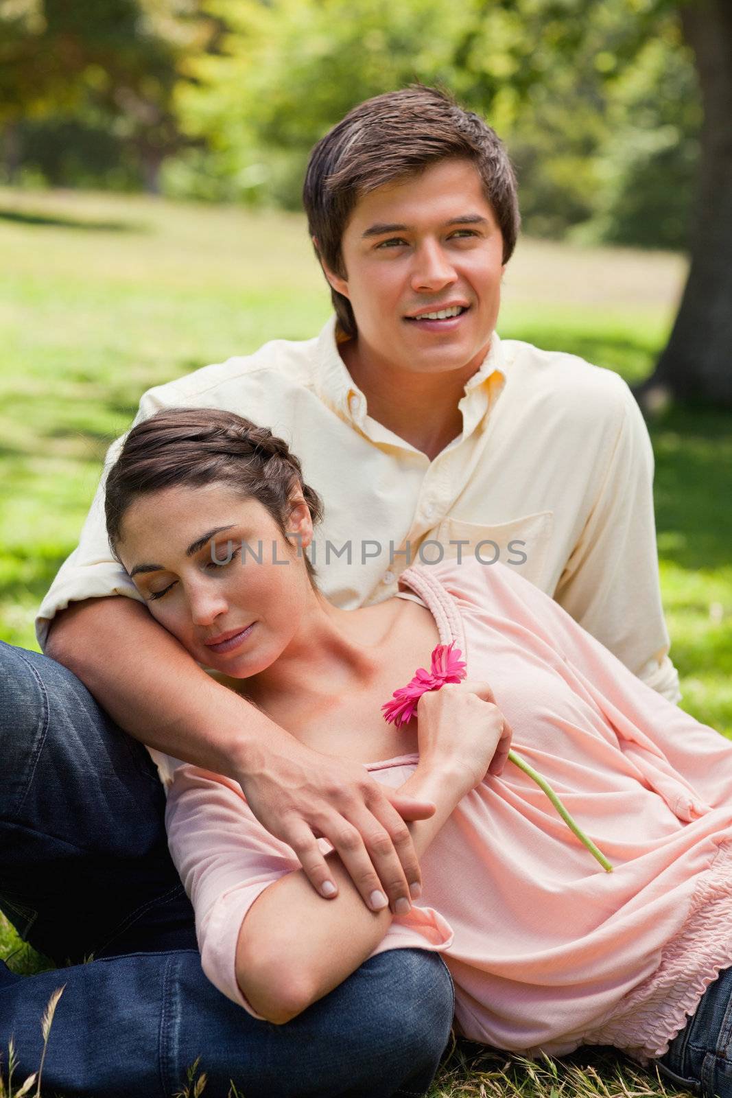 Woman with her eyes closed while holding a flower as she rests against her friend who is sitting in the grass