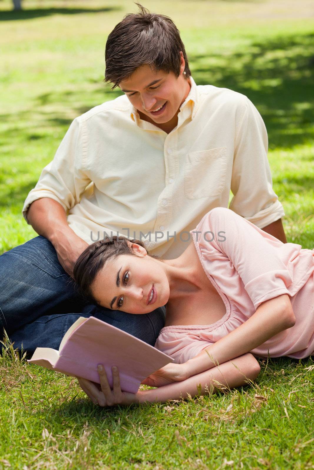 Woman lying on her side while reading a book as her friend looks at her as he sits on the grass