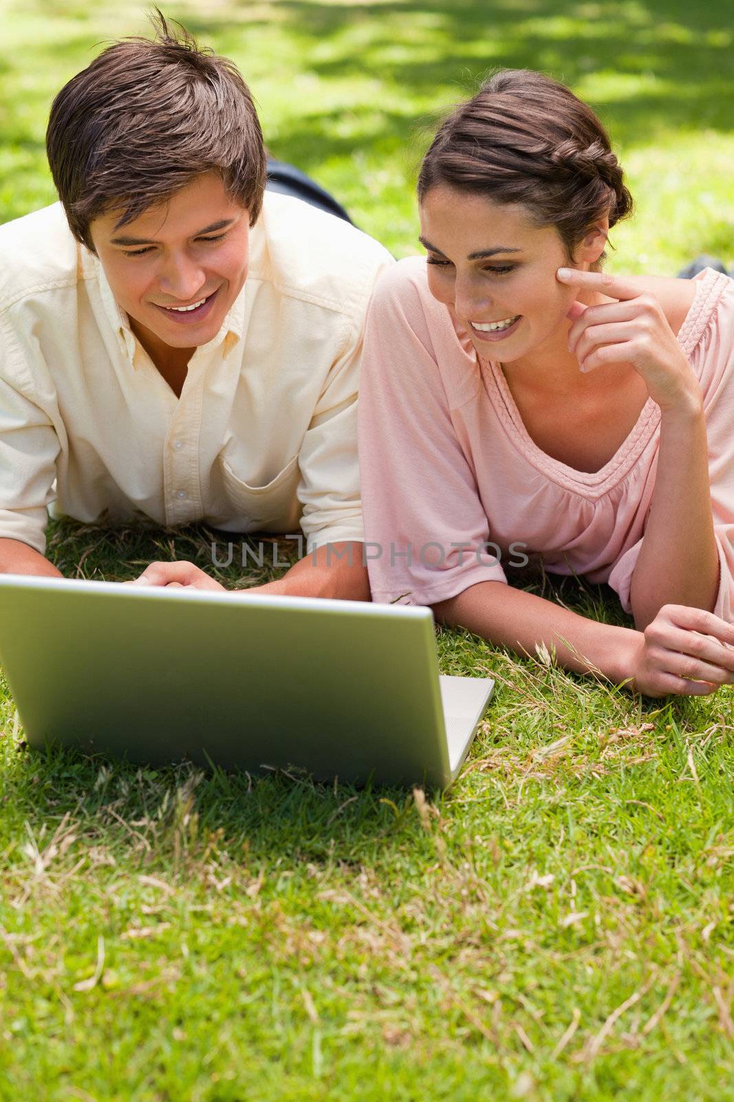 Two friends smiling while using a laptop together as they lie down in the grass