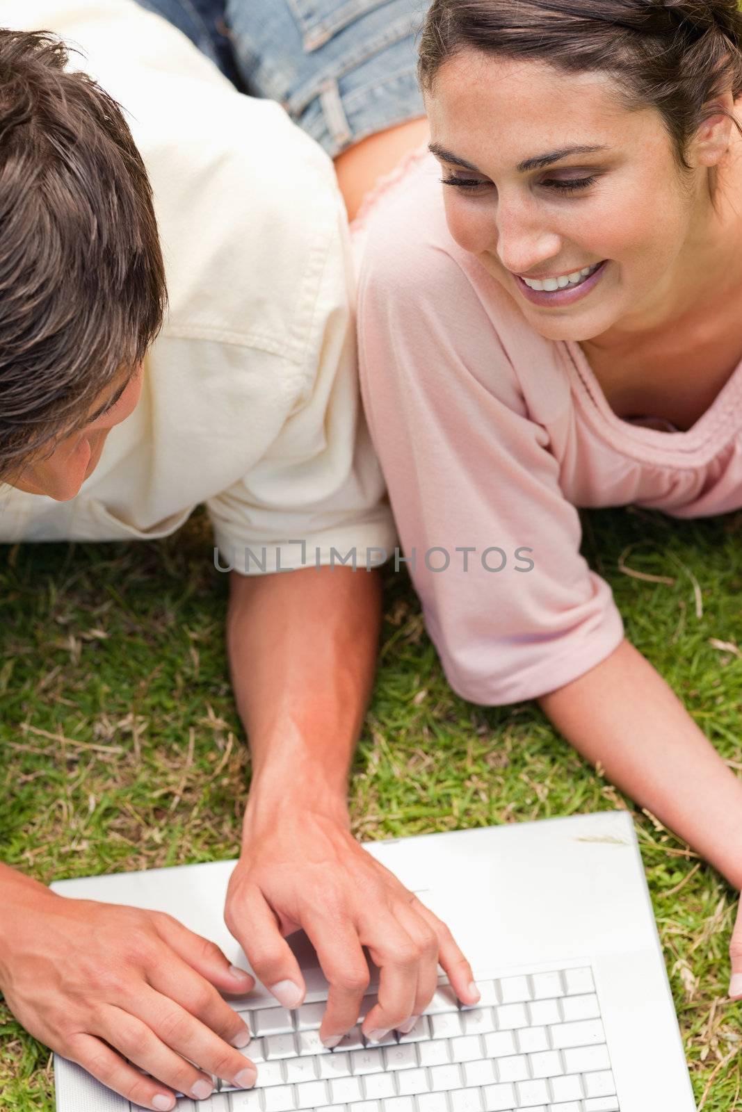 Two friends smiling while lying in the grass together and using the laptop which is in front of them