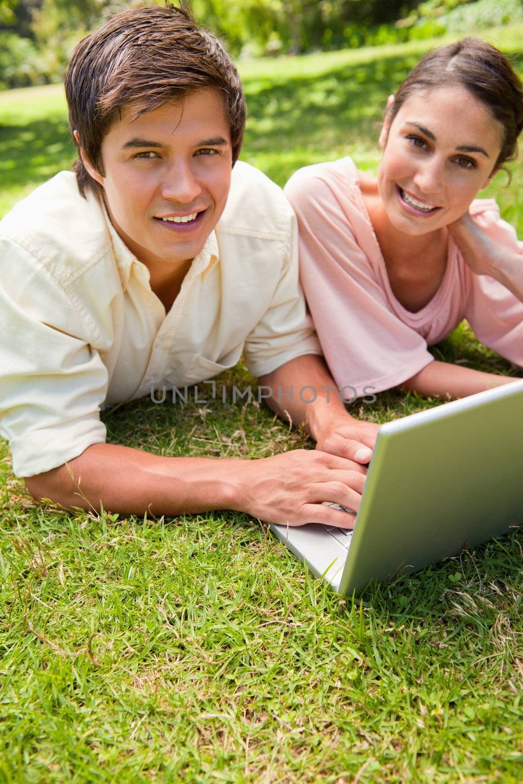 Two friends with happy expressions looking towards the side while using a laptop as they they lie together on the grass
