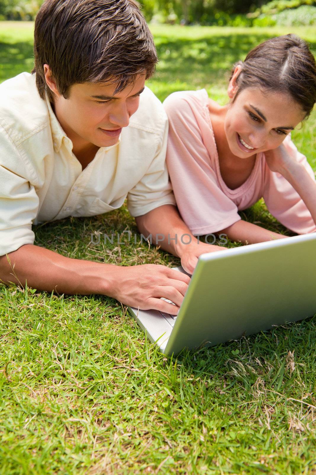 Two friends smiling as they use a laptop together while lying down on the grass