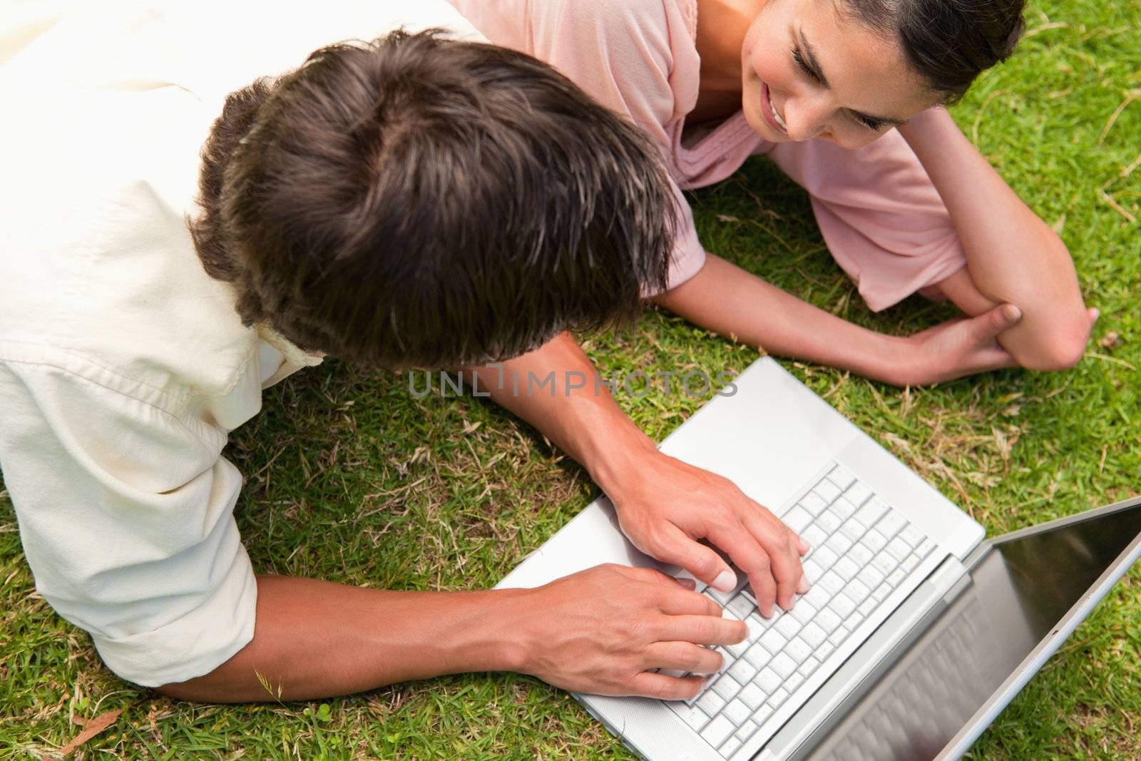Elevated view of two friends smiling while using a laptop together as they lie down on the grass