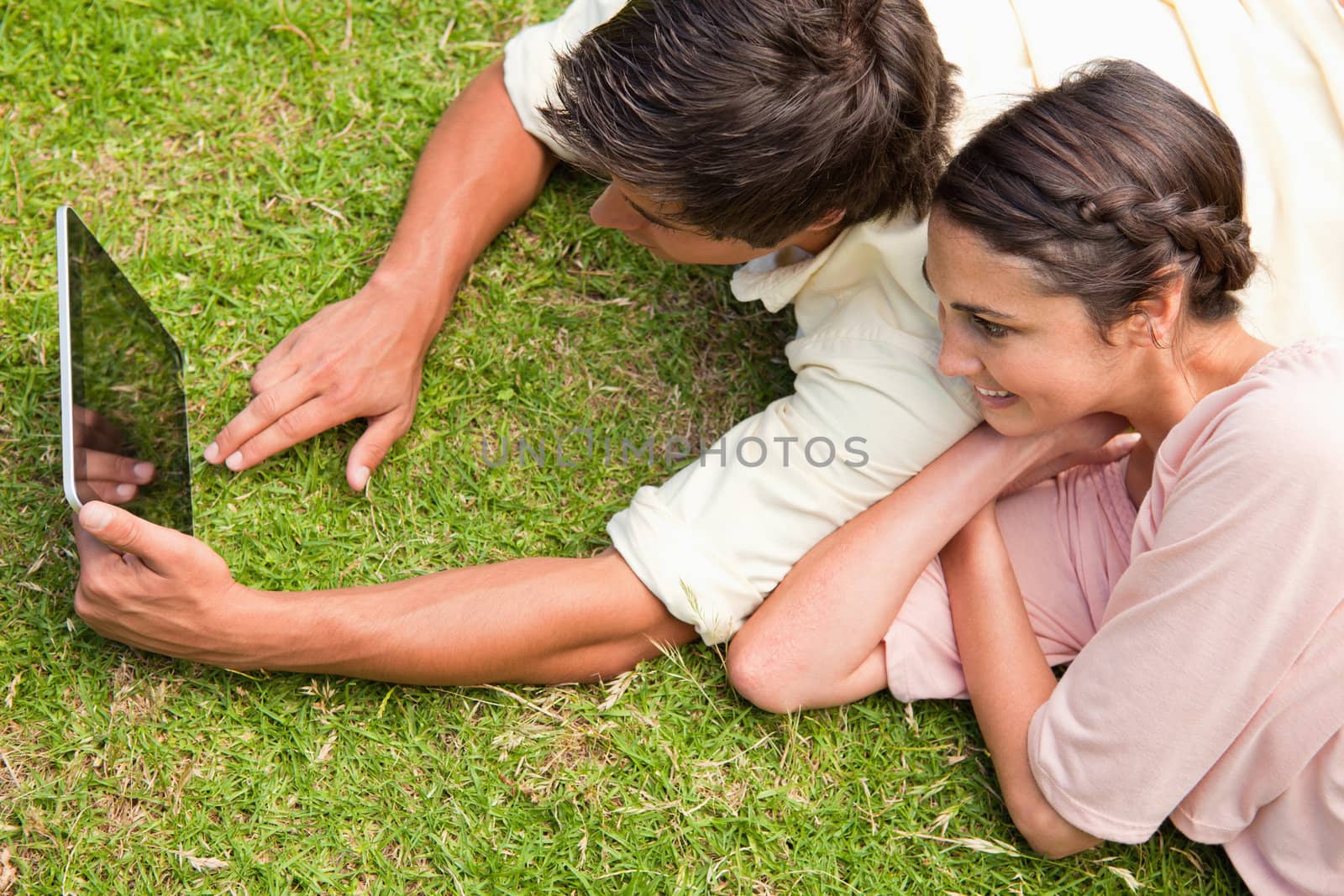 Two friends smiling as they use a tablet together while lying down on the grass