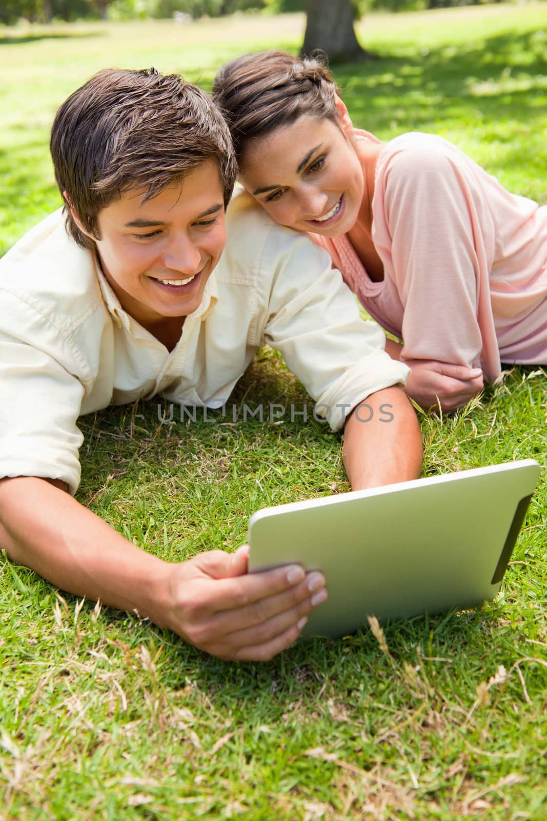 Smiling woman leans against her friend as they use a tablet together while lying down on the grass