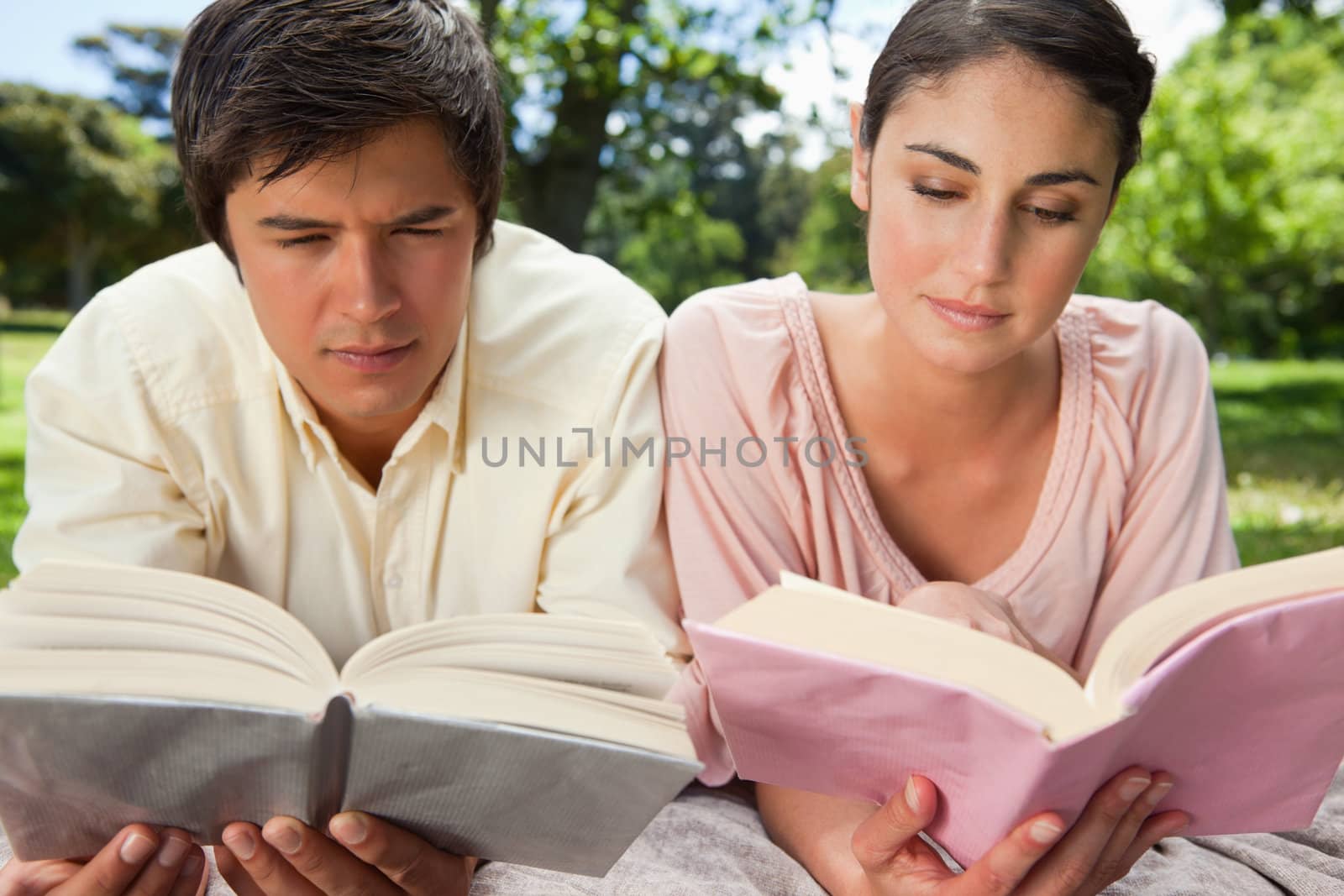 Man and a woman with a serious expression while reading books as they lie prone on a blanket in the grass