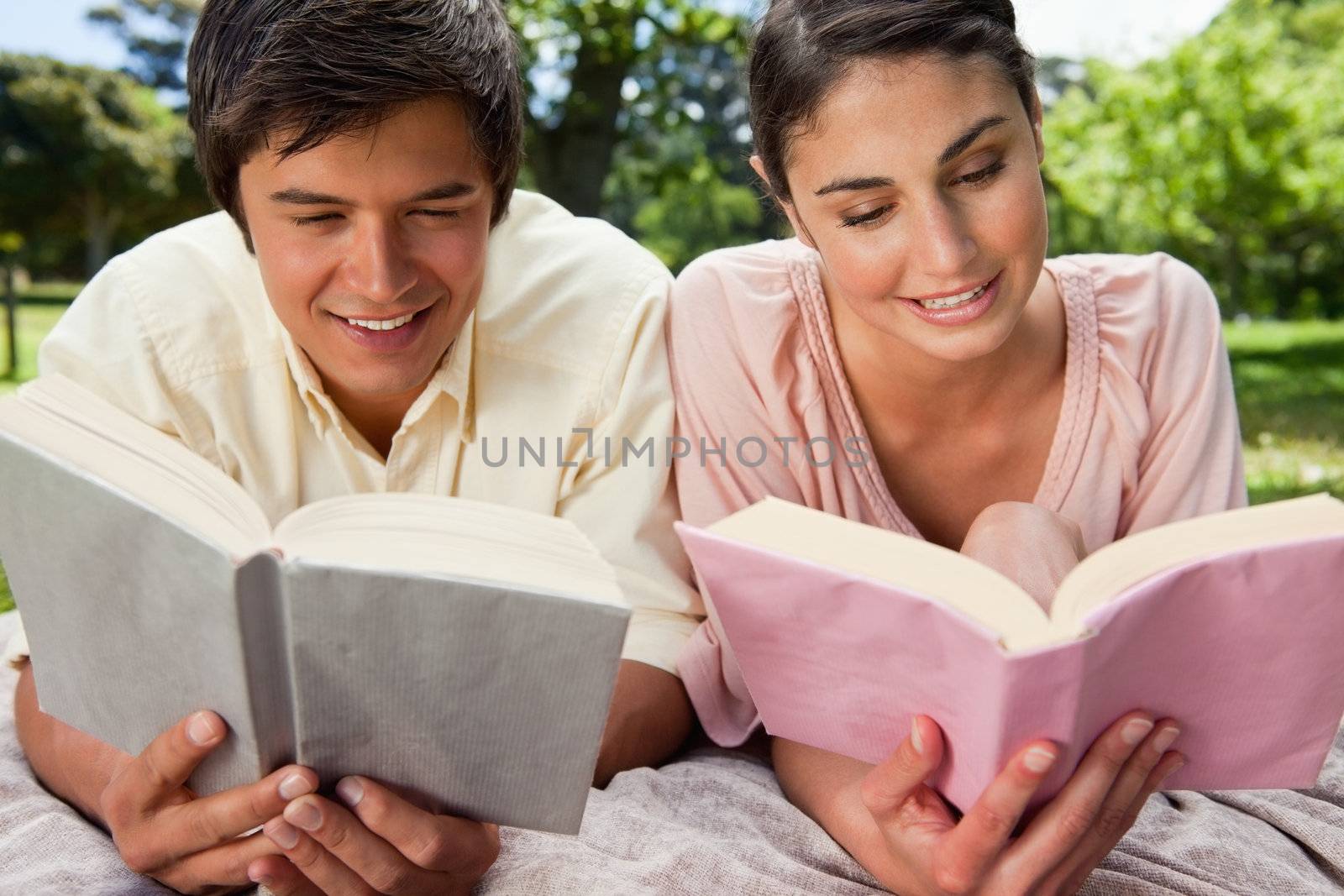Woman and a man smiling as they are reading while lying prone on a grey blanket in the grass