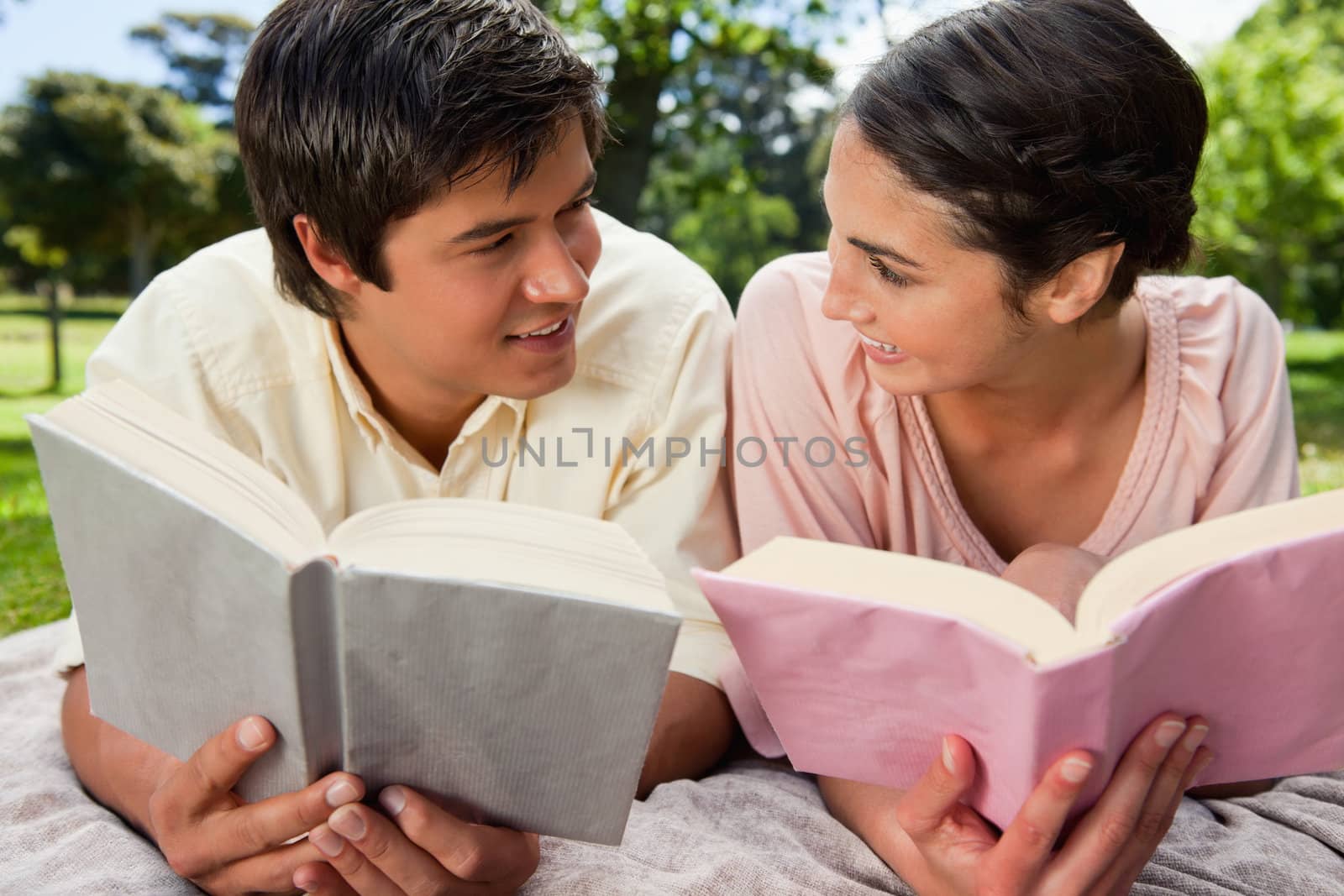 Man and a woman smiling at each other while reading books as they lie prone on a grey blanket in the grass