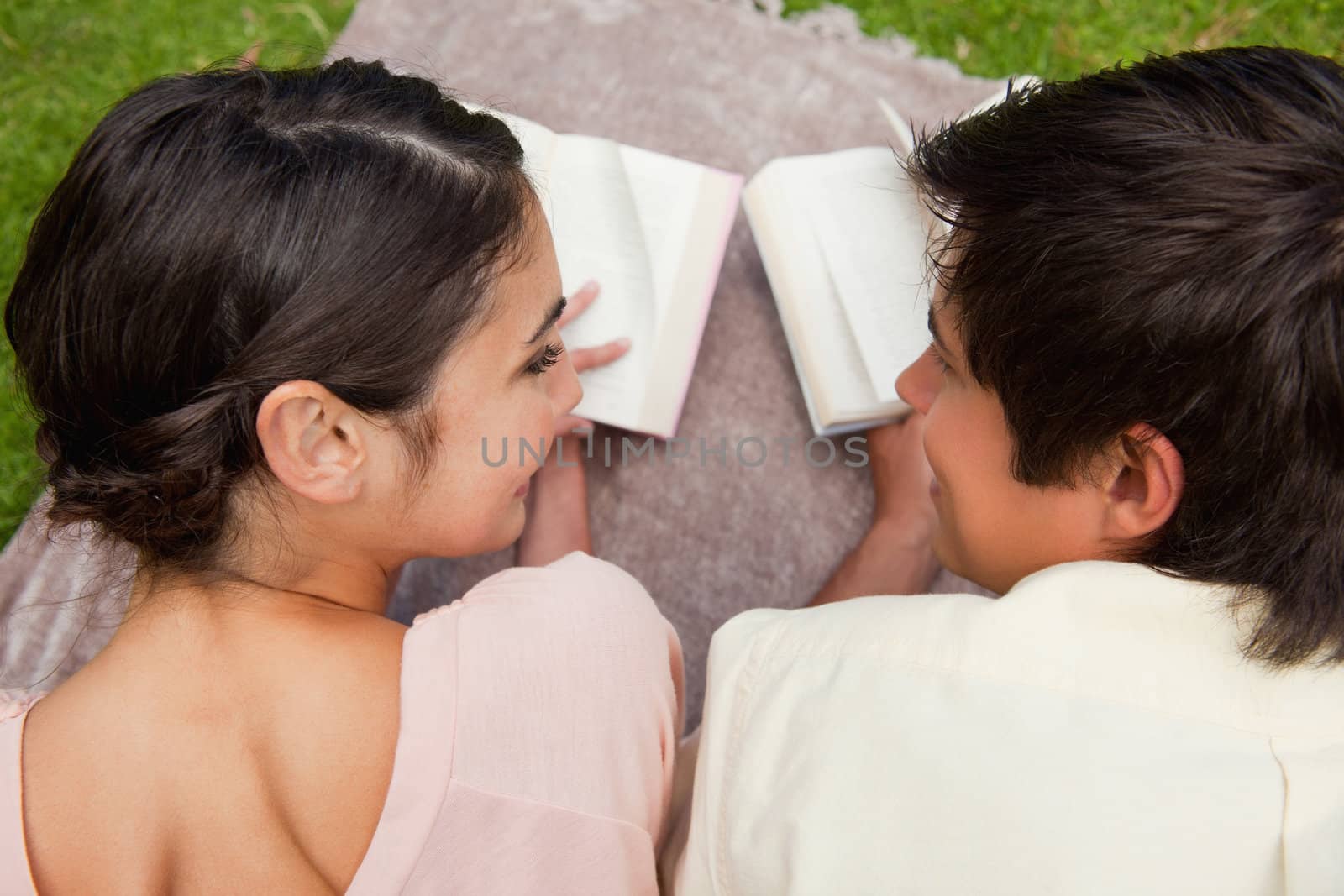 Rear view of a man and a woman looking at each other while reading books as they lie prone on a blanket in the grass