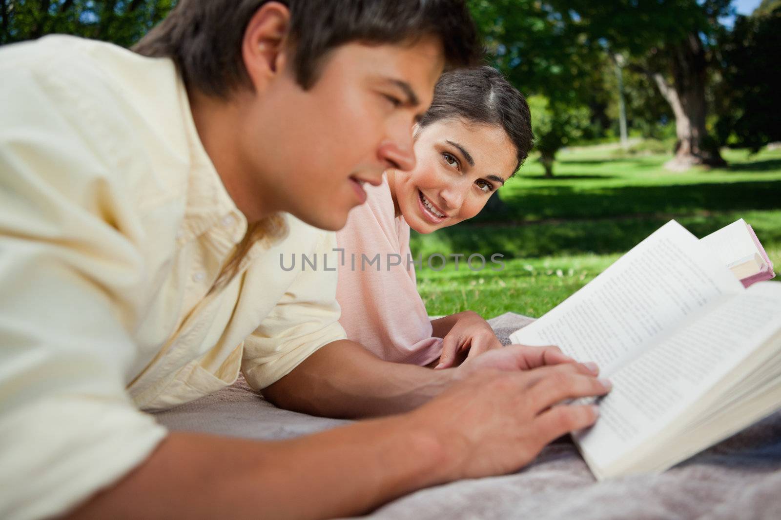 Woman looks towards the side while reading with her friend as they lie prone on a blanket in the grass with trees in the background