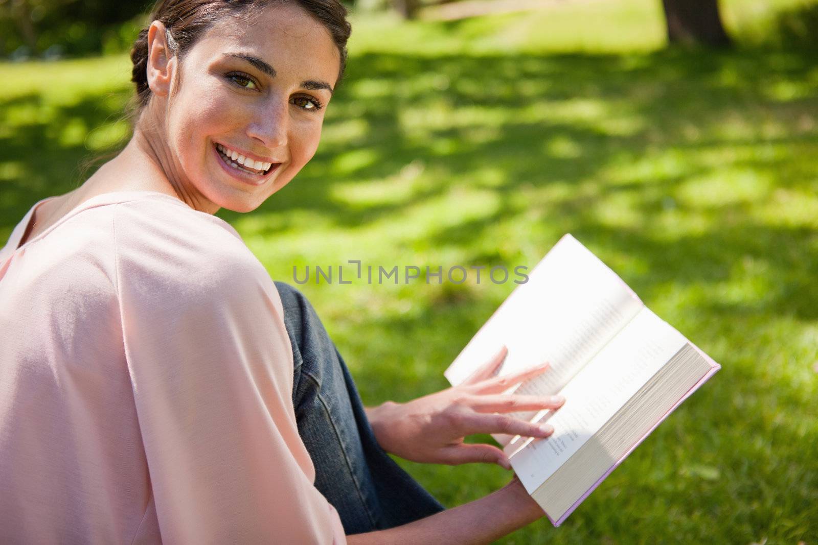 Woman looking towards her side and smiling while reading a book as she sits down in the grass