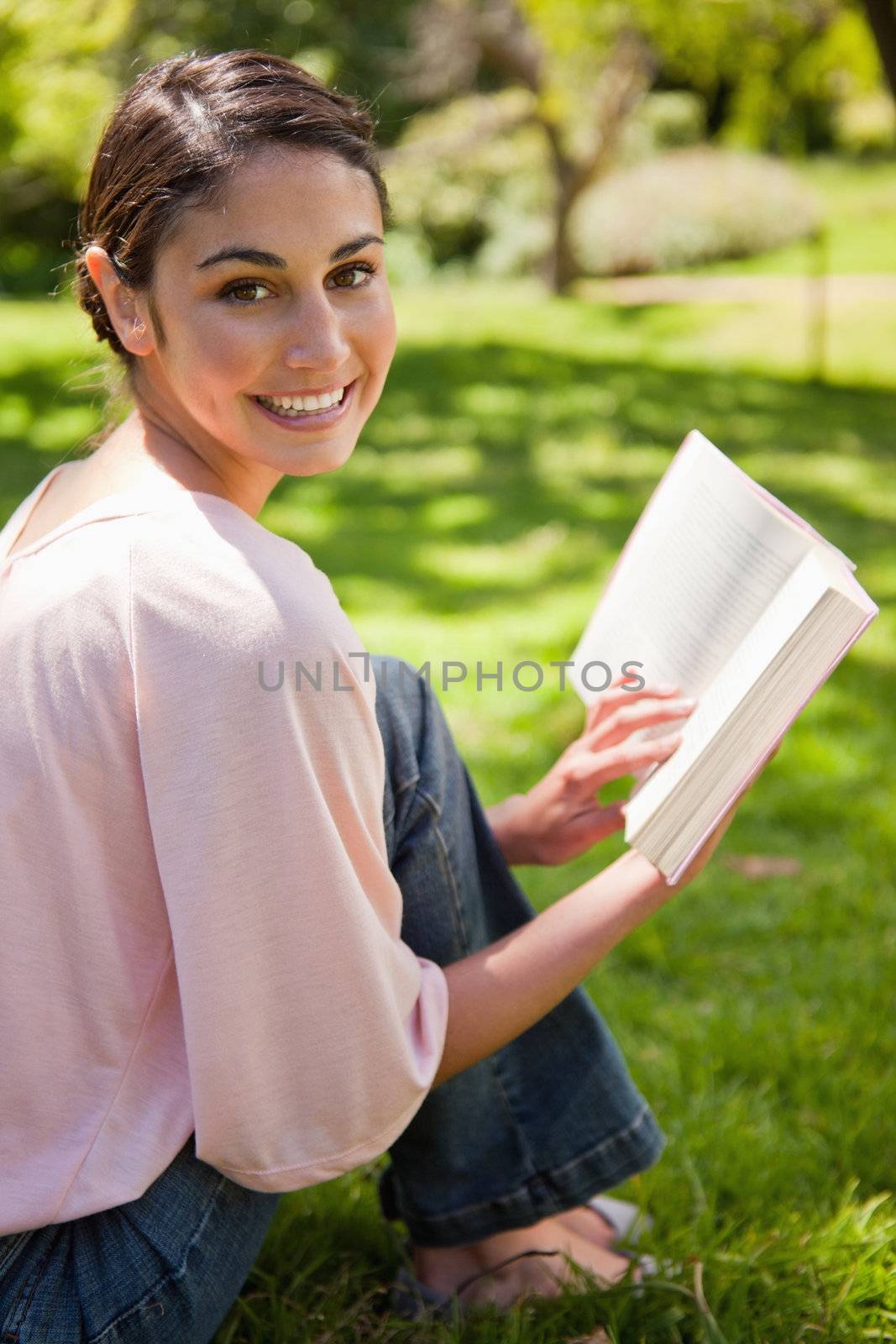 Woman looking towards her side while reading a book as she sits in a park with trees in the background