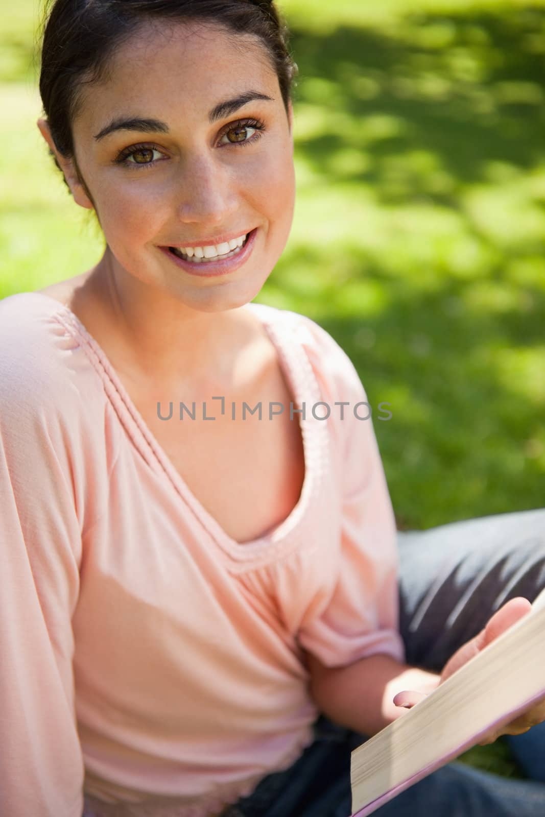 Woman looking straight in front of her as she reads a book while sitting down in grass on a sunny day