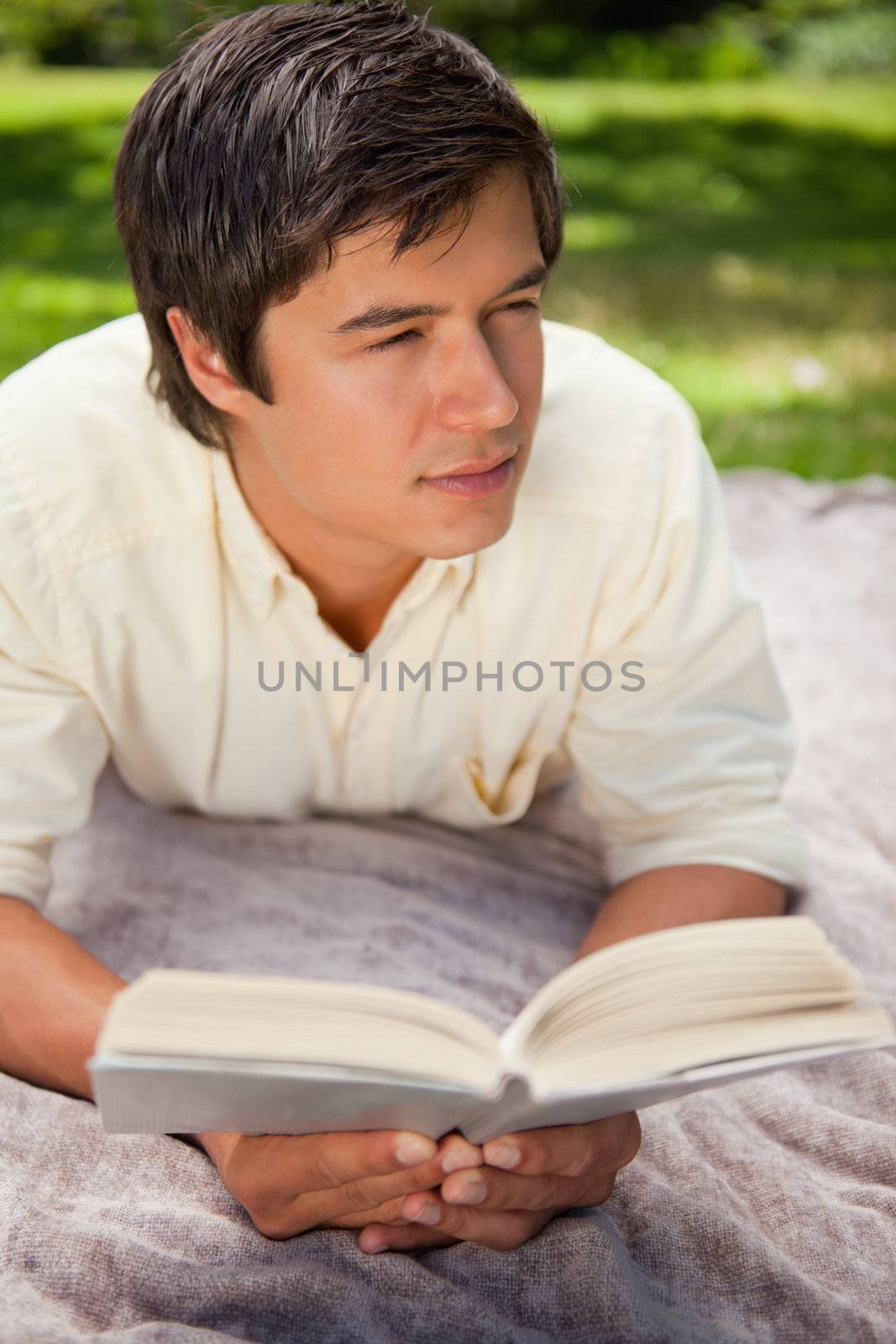 Man looking towards the side while reading a book as he lies on a grey blanket in the grass