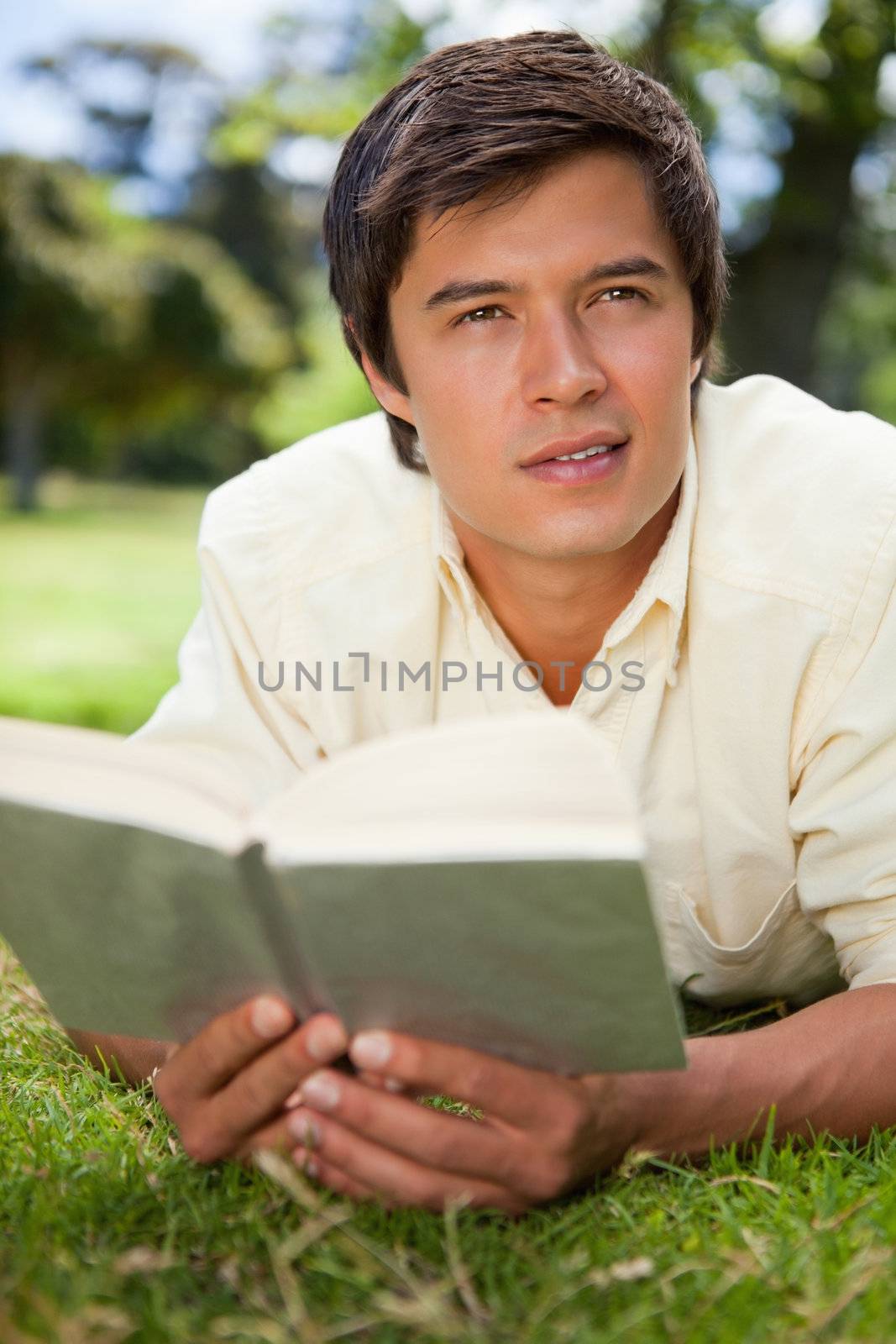 Man looking into the distance while reading a book as he lies prone in the grass