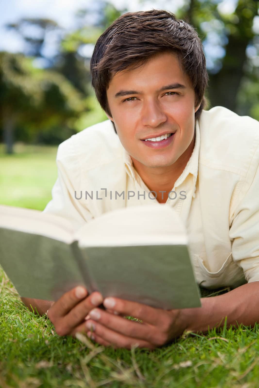 Man looking straight ahead of him and smiling while reading a book as he lies on grass
